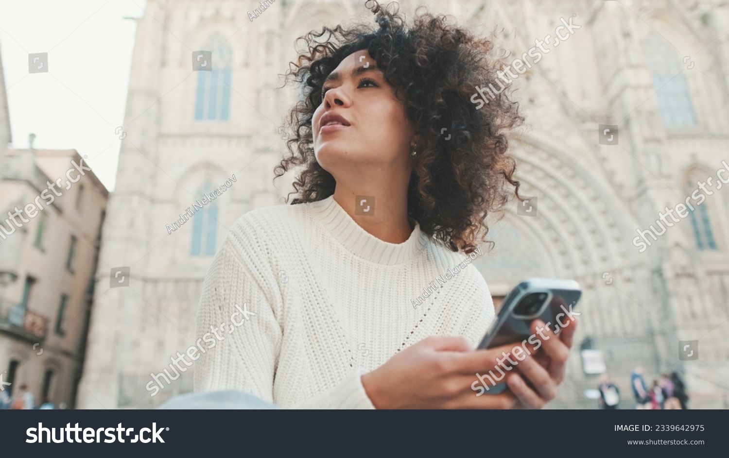 Young woman looks around. Happy girl uses mobile phone on old city background #2339642975