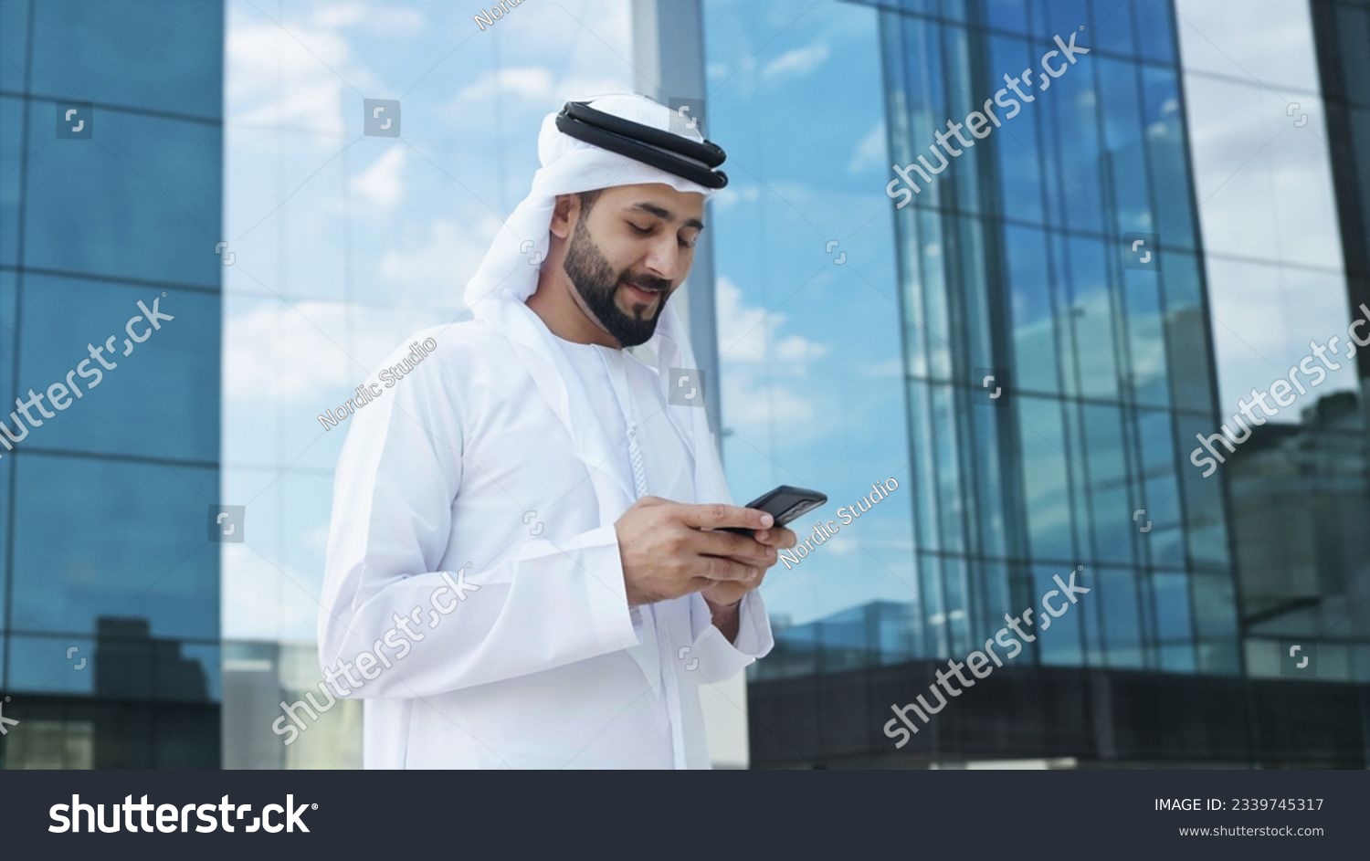 Arab businessman with high-rise buildings with glass windows as background. Arabic Man in Kandura Thobe Thawb for Middle Eastern corporate finance banking concept #2339745317