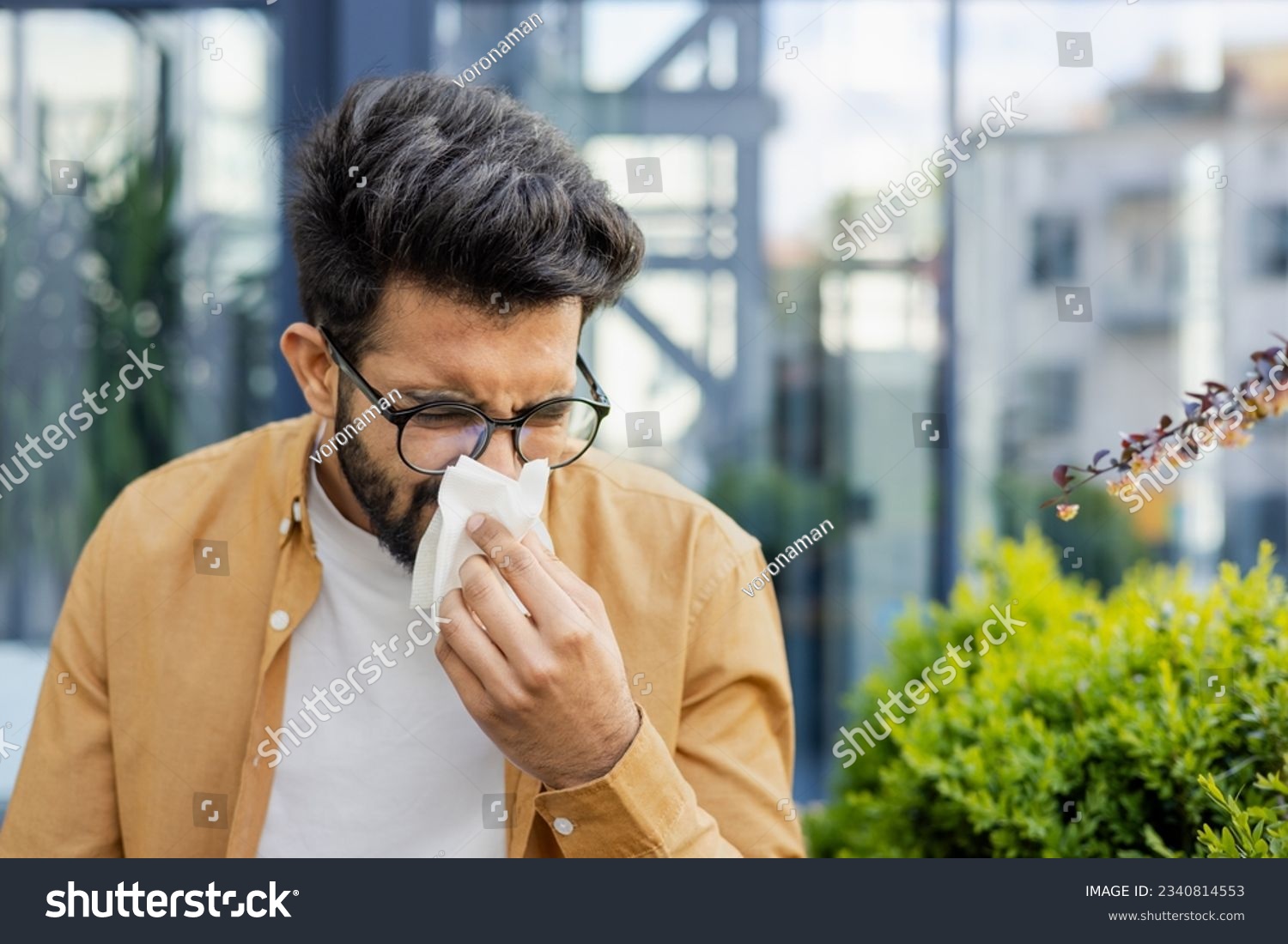 Young man sneezing and having a runny nose allergy sitting on a bench in the daytime outside an office building, hispanic businessman sick with a tissue near his nose. #2340814553