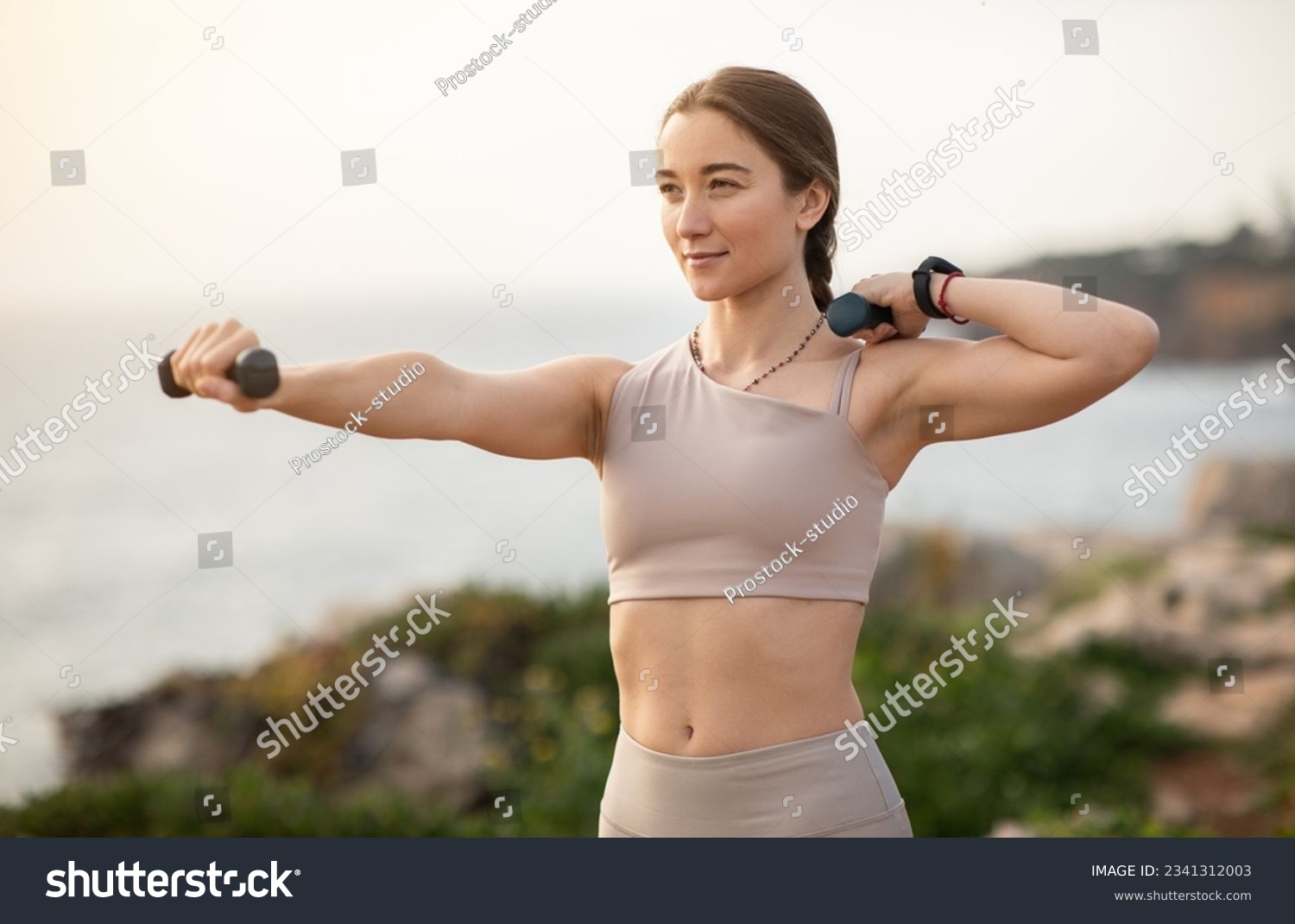 Glad confident strong millennial caucasian woman athlete in sportswear doing arm exercises with dumbbells, enjoy workout, body care alone at sea beach. Sports, fitness outdoor #2341312003