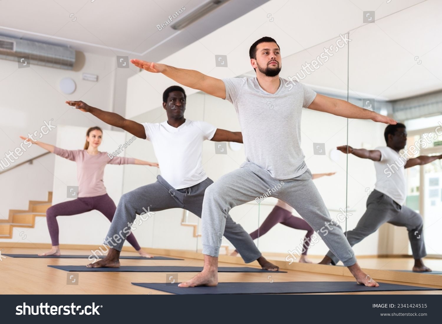 Caucasian man practising warrior II pose with people during group yoga training. #2341424515