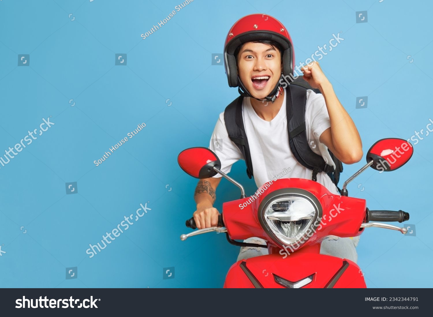 Asian happy guy in red helmet riding red motorbike and holds one fist up, posing on blue backdrop with wide smile, lifestyle concept, copy space #2342344791