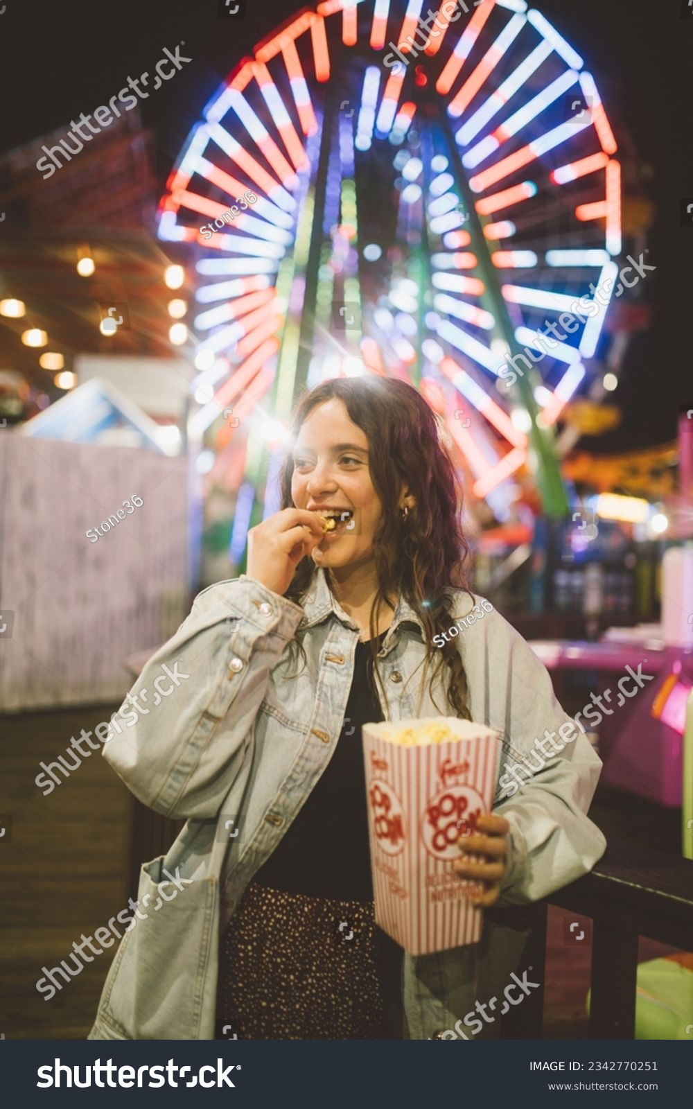 A young lady in a good mood with a popcorn cup in the night at luna park in Santa Monica in front of unfocused illuminations of a Ferris wheel. Santa Monica. California. #2342770251