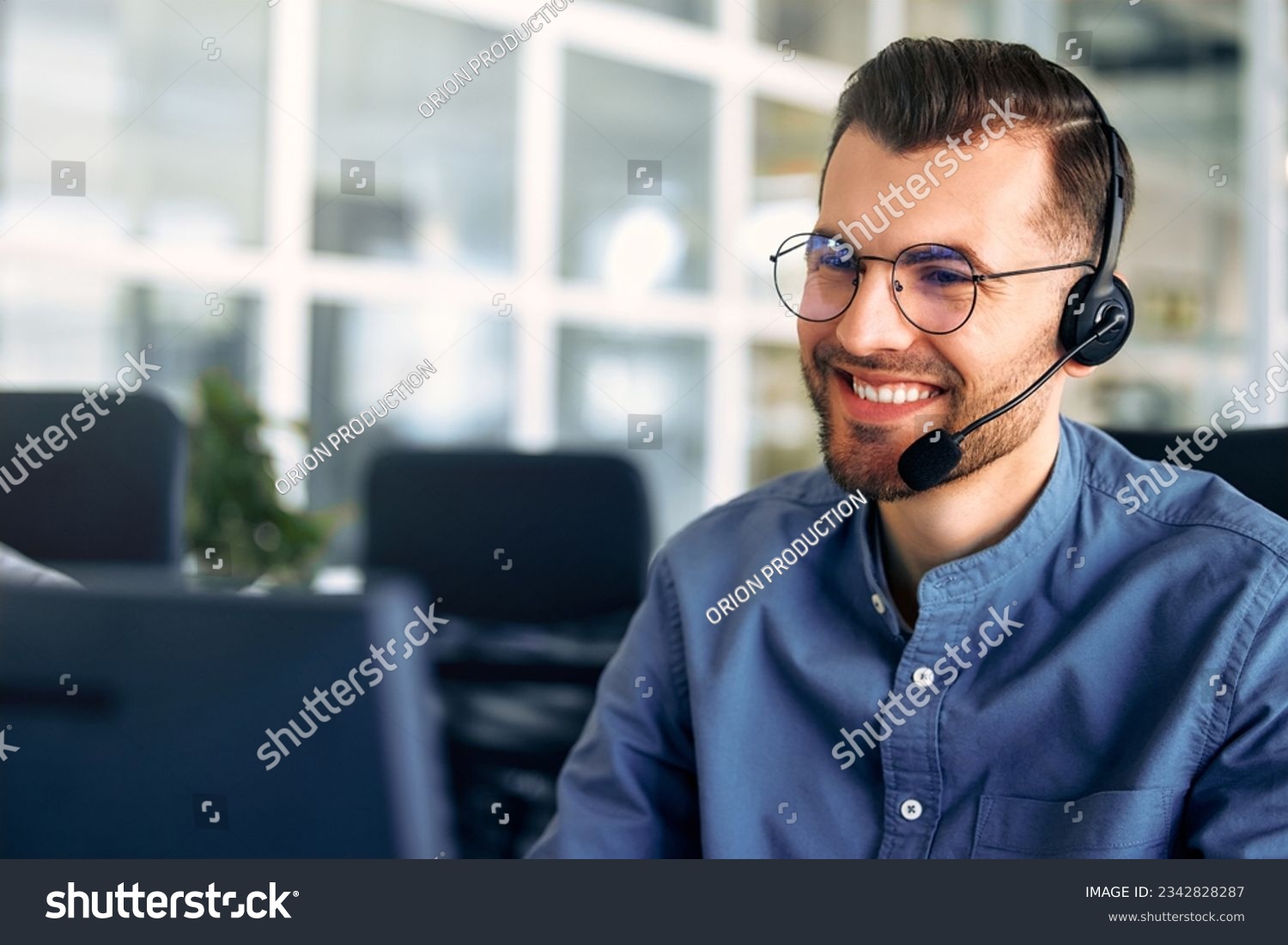 Technical support operator working with headset in office. Smiling handsome man working as call centre operator, speaking to customer. Happy businessman working remotely while doing video conference. #2342828287