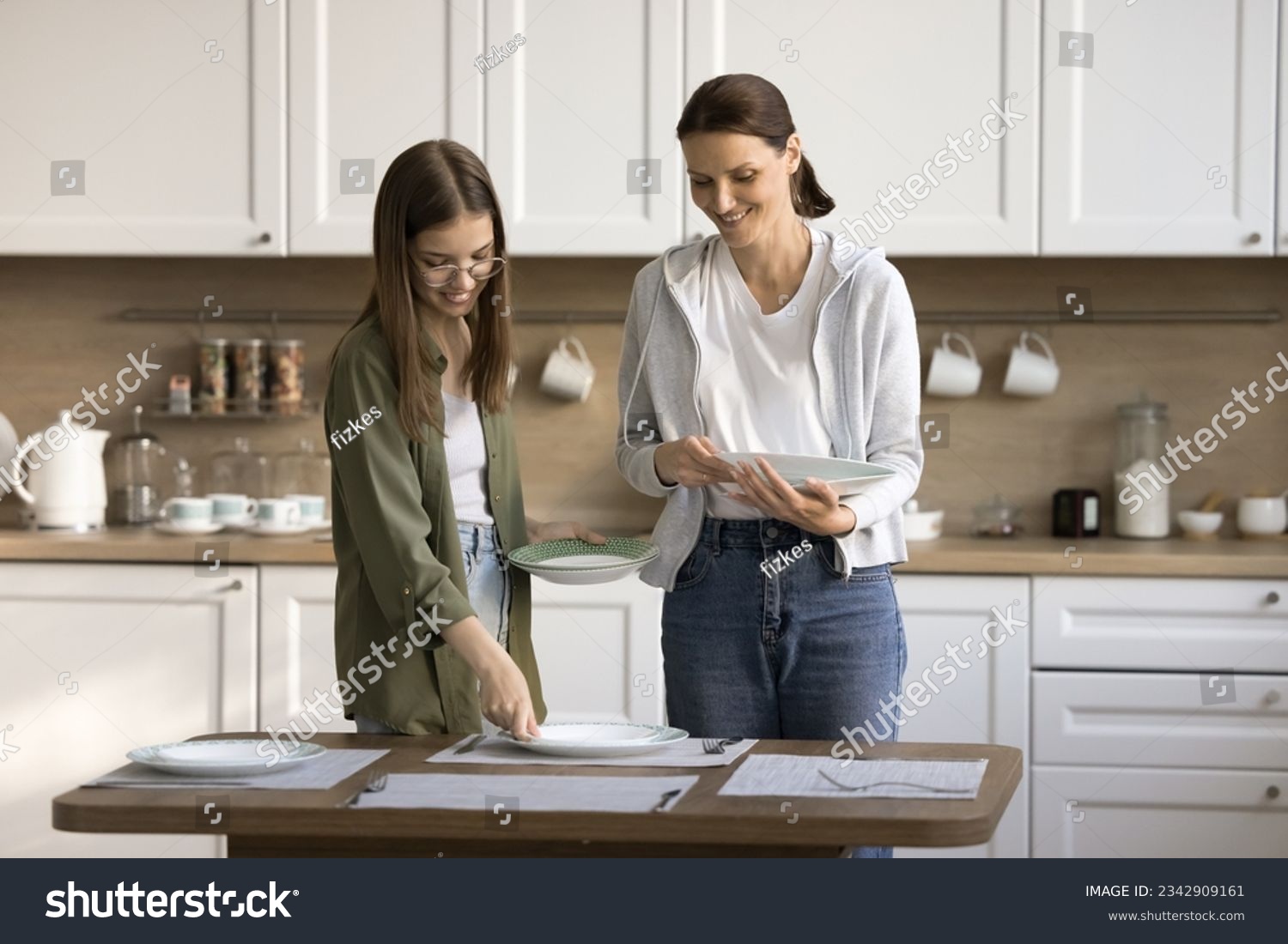 Happy mother and teen daughter child decorating table for dinner, placing dich, plates, talking, chatting, smiling, laughing, enjoying household activities. Kid helping mom with domestic chores #2342909161