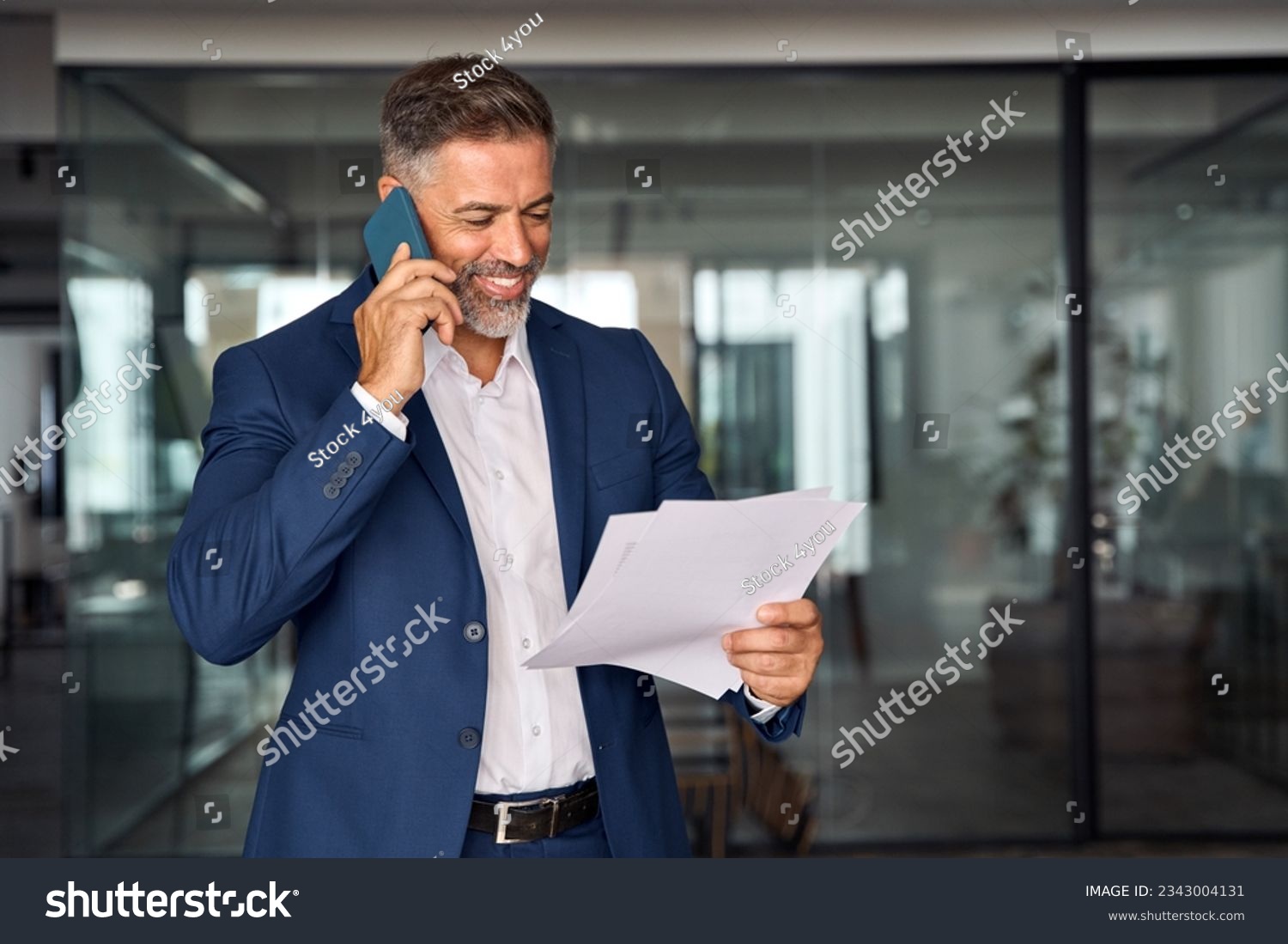 Middle aged Latin or Indian businessman walking, having call on smartphone with business partner and looking at documents. Mature Hispanic man talking by mobile cellphone at work in office, copy space #2343004131