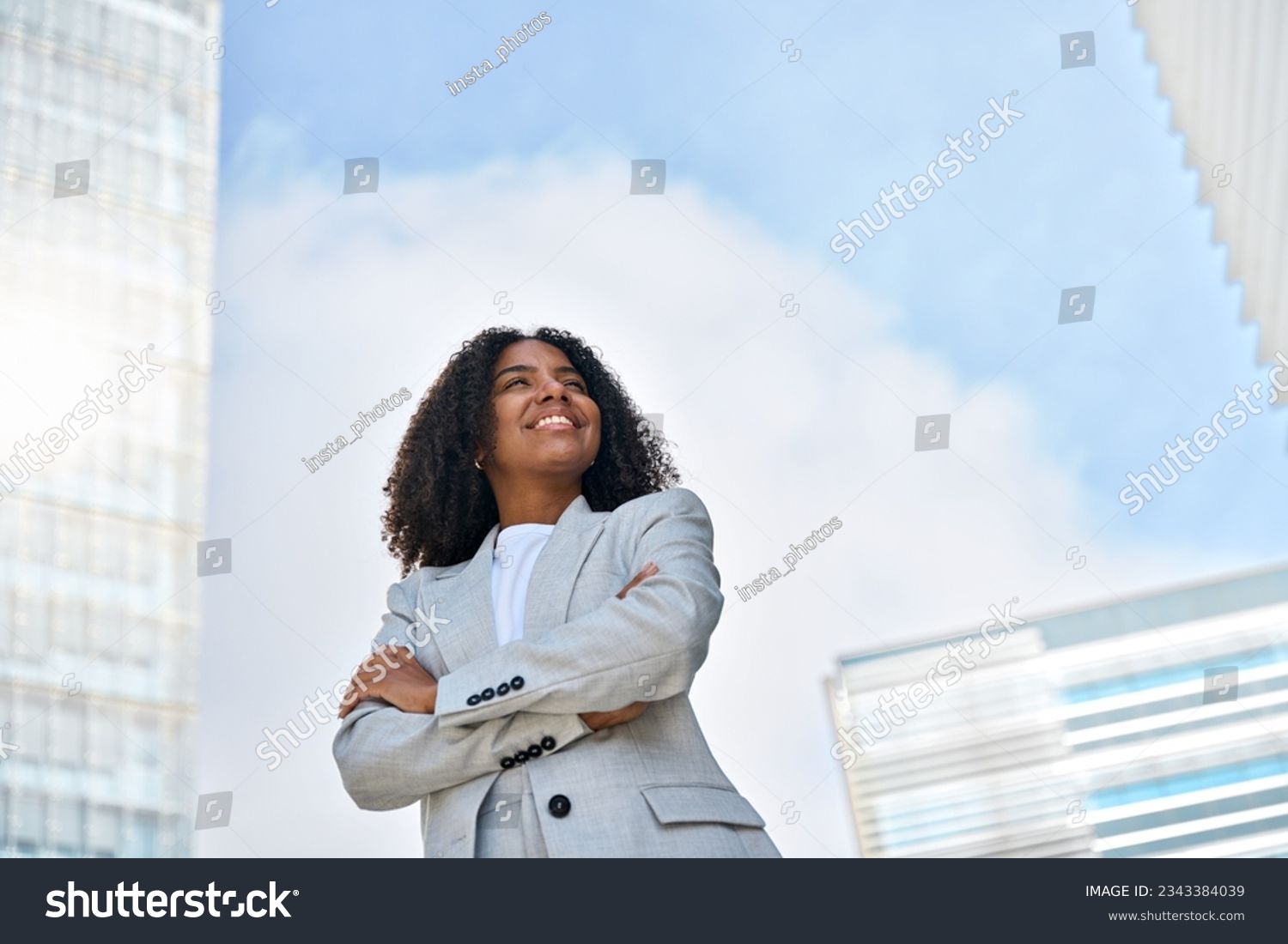 Happy smiling confident young African American professional business woman leader wearing suit standing in big city on street arms crossed thinking of success, dreaming, feeling proud outdoors. #2343384039