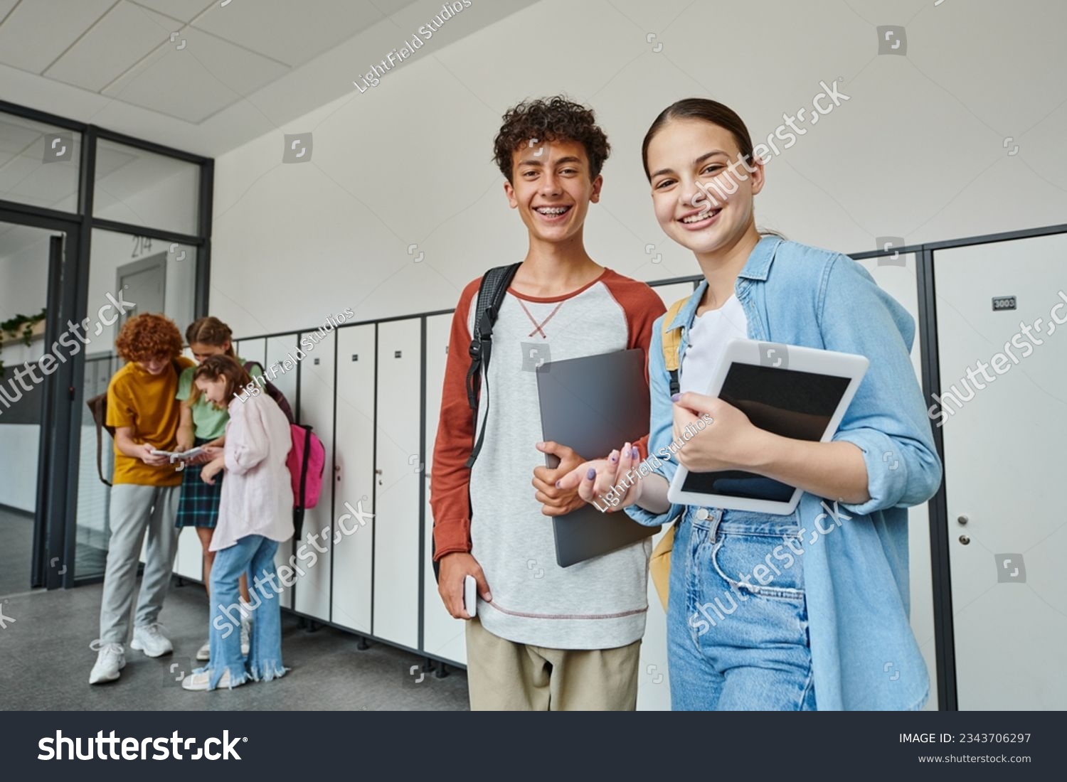 happy teen classmates holding devices and looking at camera in school hallway, adolescent students #2343706297