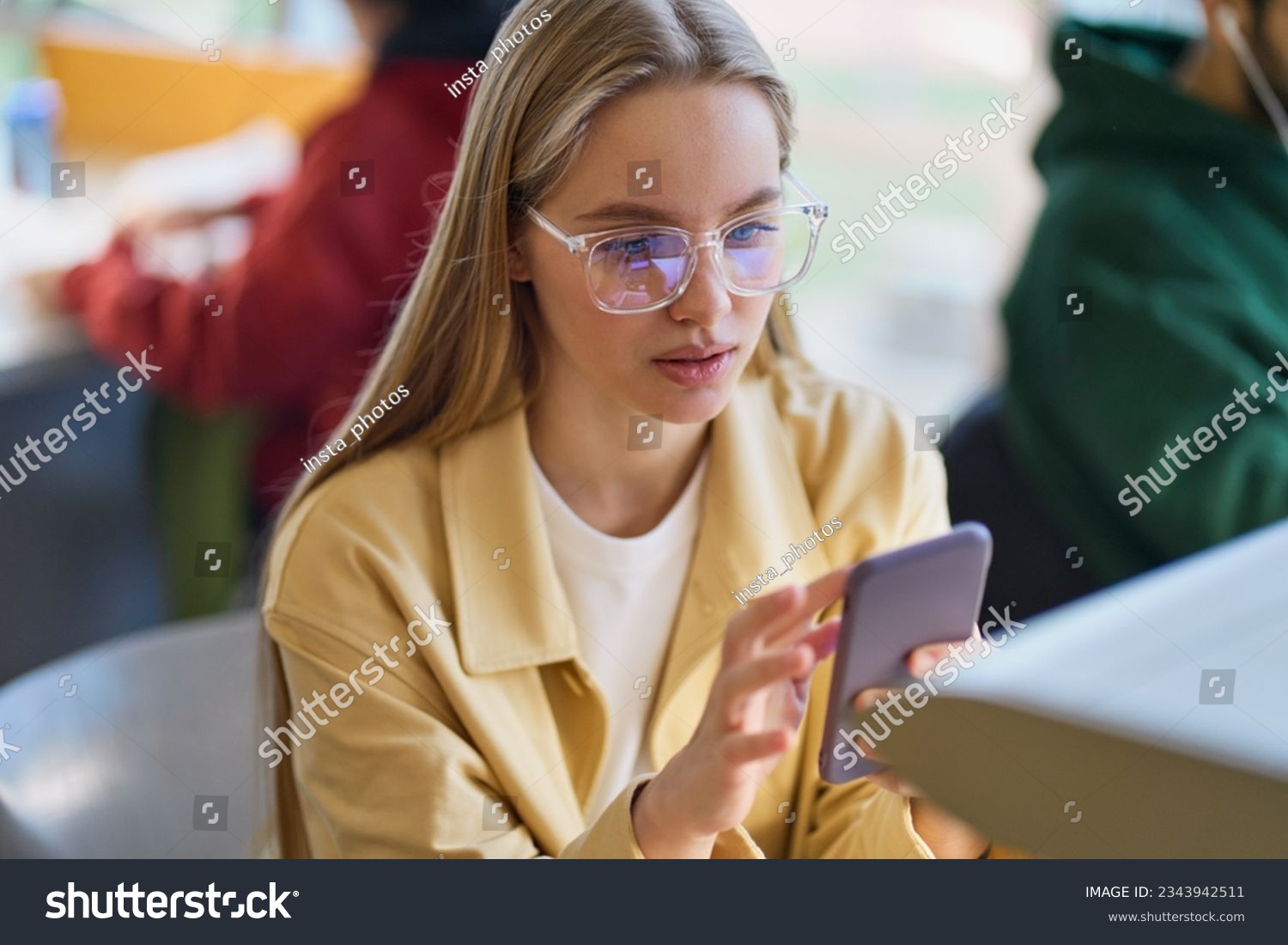 Teen girl gen z student using mobile phone looking at smartphone sitting at desk in university college campus classroom. Young blonde woman holding cellphone modern tech in university. #2343942511