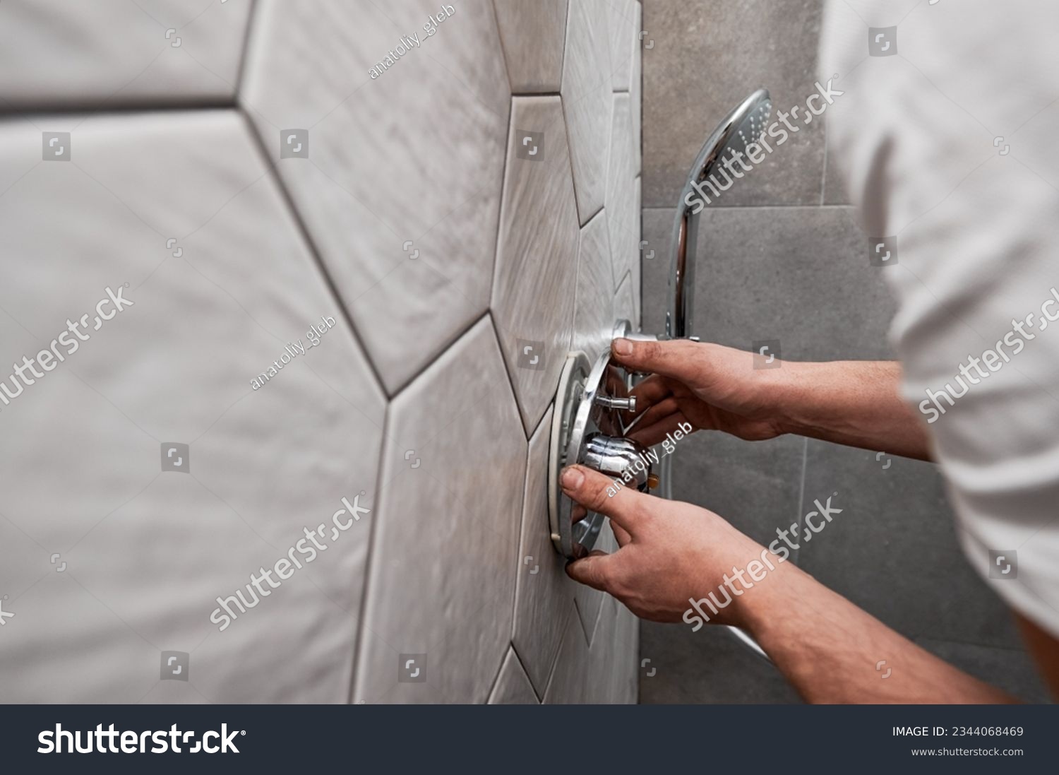 Close up of man standing by the wall with ceramic tile and installing shower faucet with metal handle in apartment. Male plumber working on bathroom renovation at home. Plumbing works concept. #2344068469