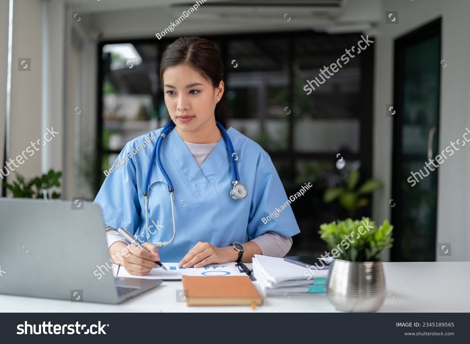 A professional and focused Asian female doctor in scrubs is working and reading medical research on her laptop in her office at a hospital. #2345189565
