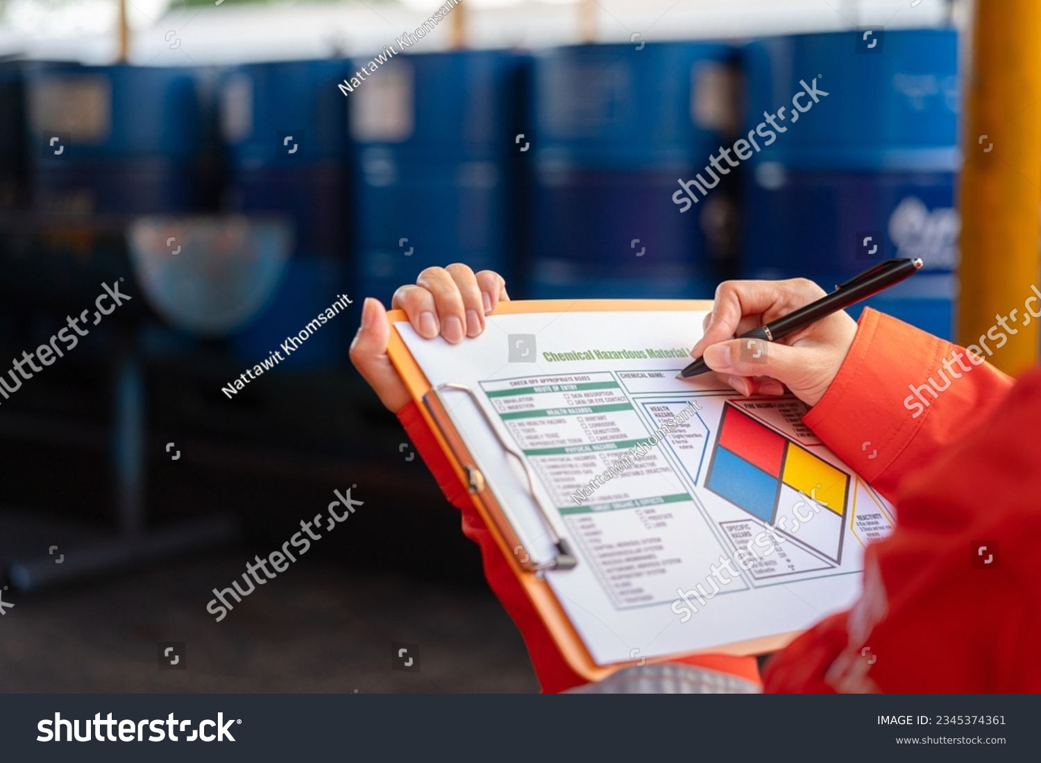 A safety officer is checking on the hazardous material checklist form with chemical storage area at the factory as background. Industrial safety working scene, selective focus. #2345374361