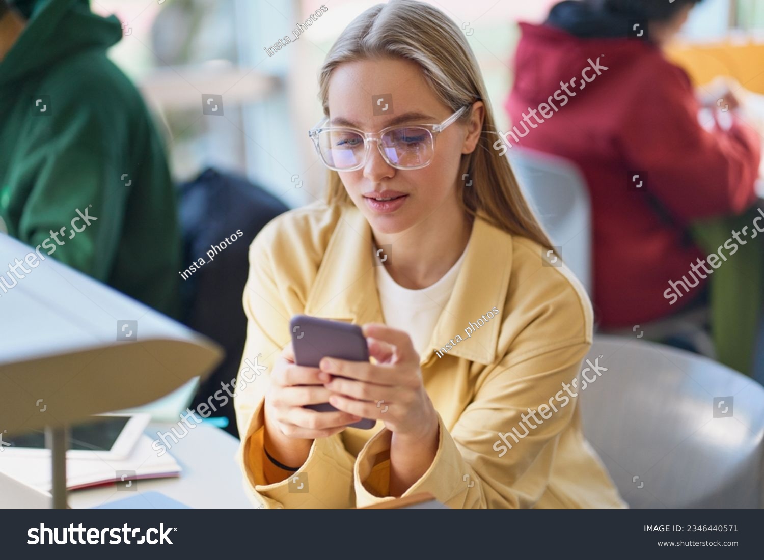 Girl student using mobile phone looking at smartphone sitting at desk in university college campus classroom. Young blonde woman holding cellphone modern tech in university advertising apps. #2346440571