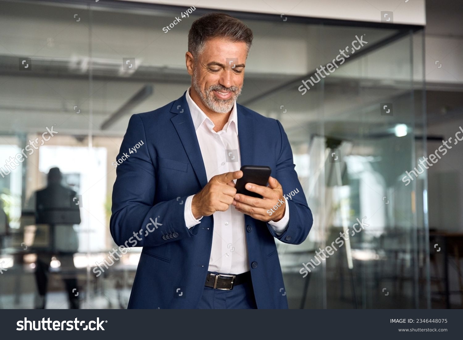 Smiling mature Latin or Indian businessman holding smartphone in office. Middle aged manager using cell phone mobile app. Digital technology application and solutions for business success development. #2346448075