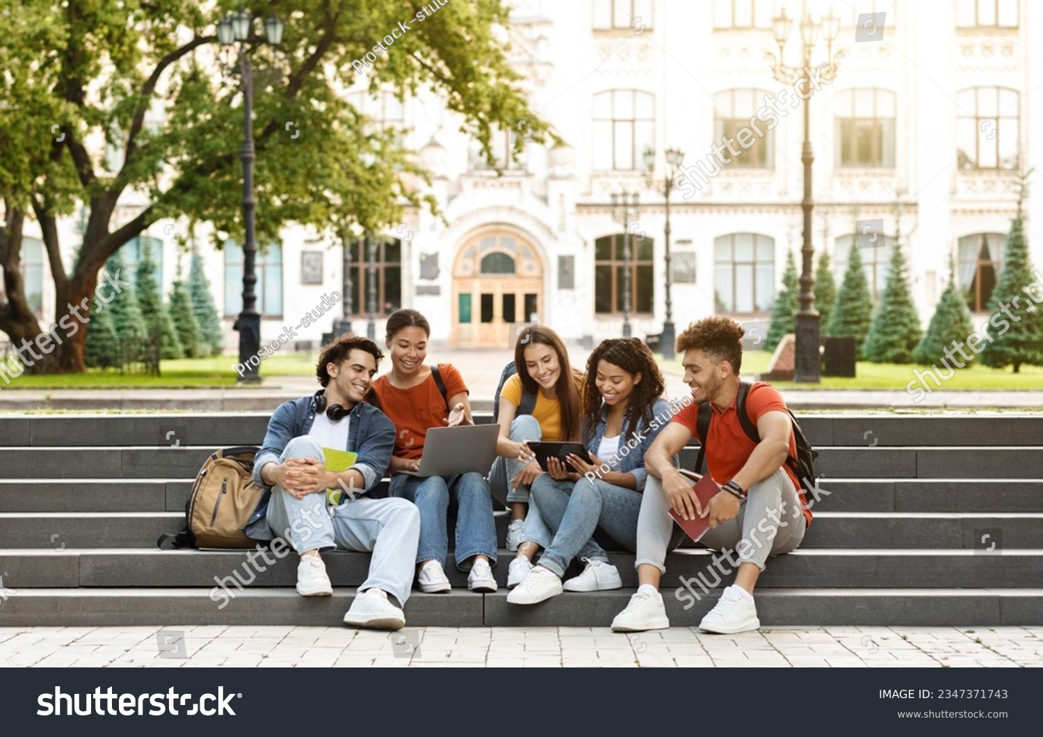 Group Of Students With Digital Tablet And Laptop Study Together Outdoors, Happy Multiethnic Young Friends Sitting On Stairs Near University Building, Using Modern Gadgets For Education, Copy Space #2347371743