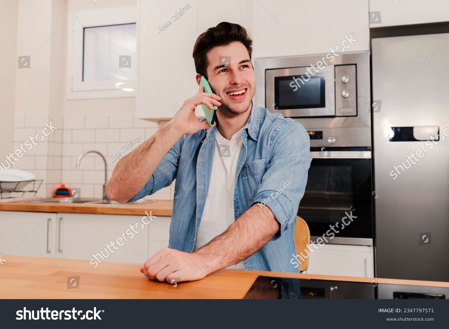 Young caucasian handsome guy having a friendly telephone call conversation sitting at home with positive expression. One young man smiling, laughing and talking by cell phone at kitchen room. High #2347797571