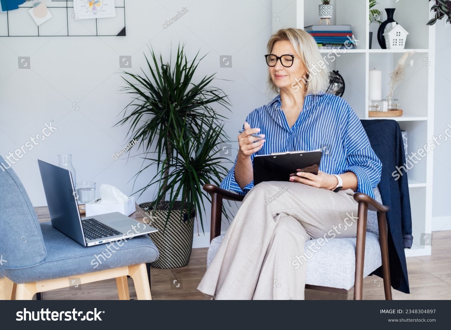 Professional Psychotherapy online. Middle aged female Psychologist having video call consultation on laptop while sitting on armchair In office. Specialist talking, counseling, helping patient online #2348304897