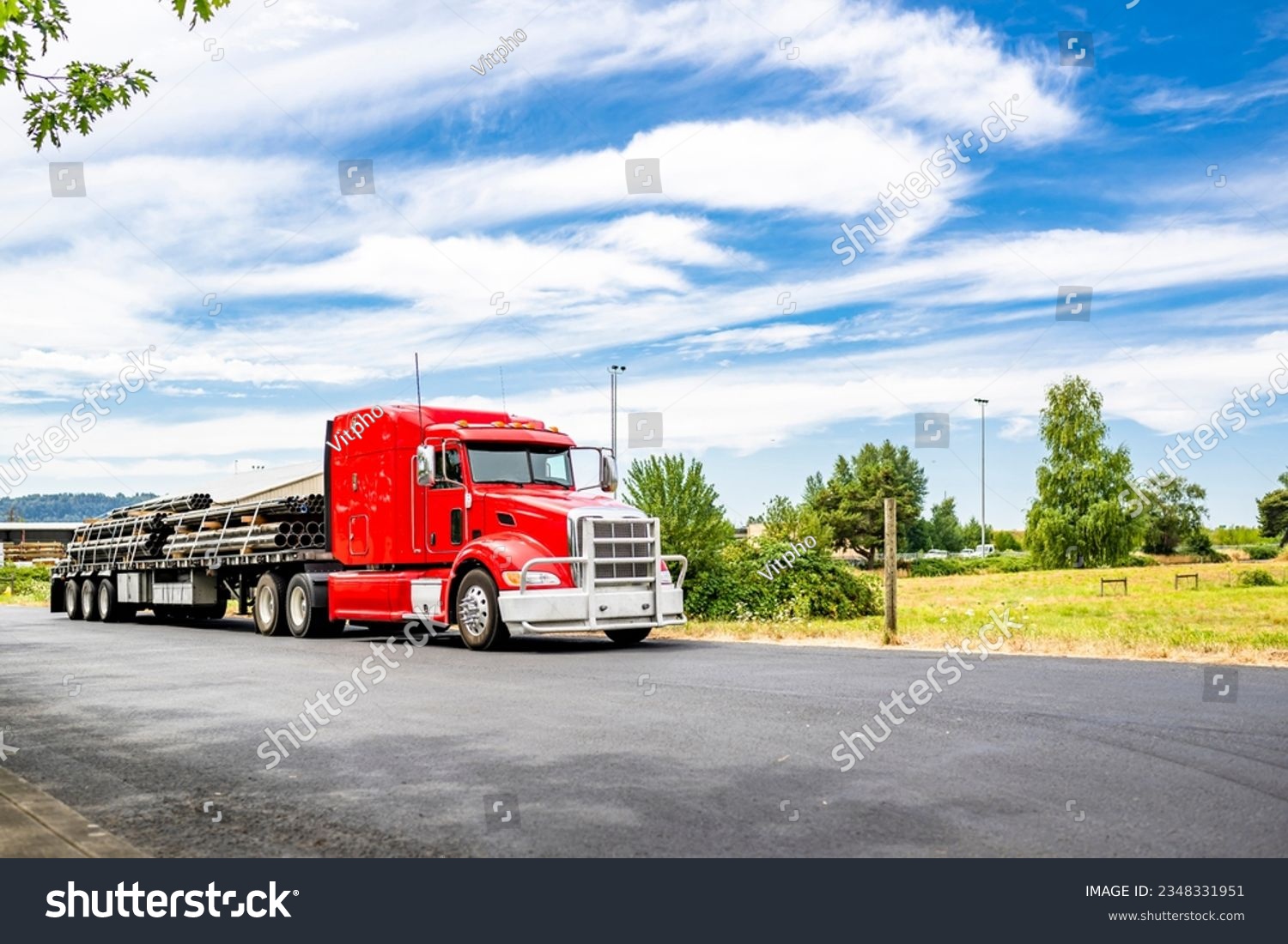 Long hauler big rig red semi truck tractor with grille guard and loaded by pipes flat bed semi trailer standing on the local road take a break for truck driver rest according to log book schedule #2348331951
