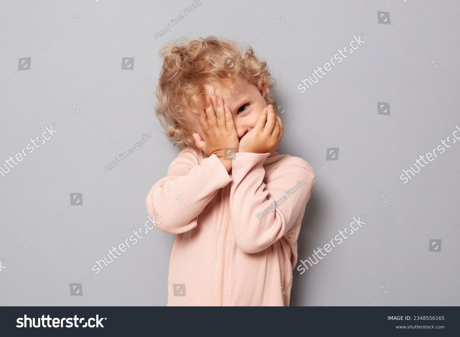 Adorable baby portrait. Shy baby girl with blonde wavy hair wearing rose shirt isolated over gray background hiding her face with arms looking through fingers to camera #2348556165