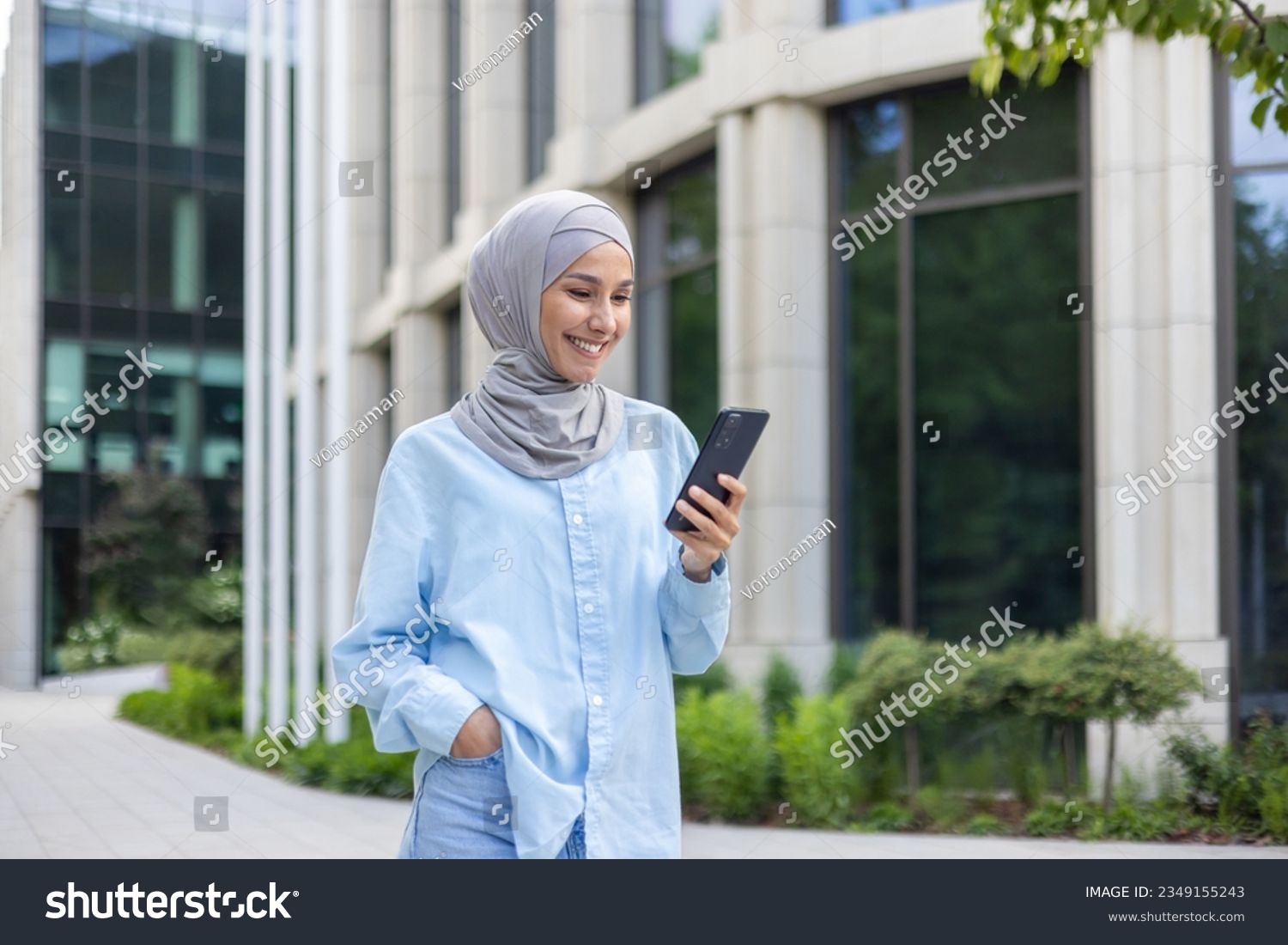 A young beautiful business woman in a hijab walks through the city, a Muslim woman holds a phone in her hands, uses an application on a smartphone, smiles, browses the Internet. #2349155243