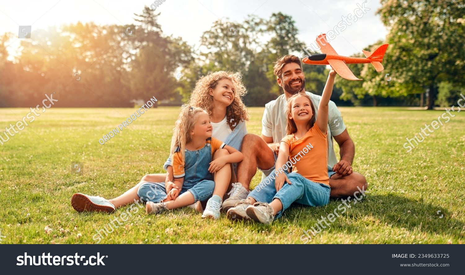 Happy young family dad, mom and two children daughters playing with an airplane sitting in a meadow on the grass in the park on a warm sunny day, having fun on a day off. #2349633725