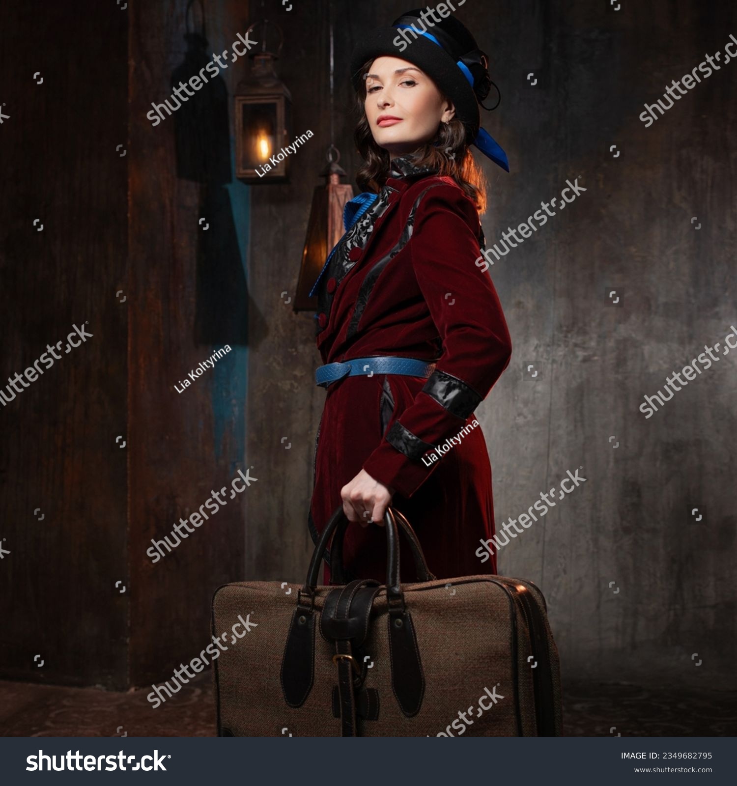 A stylish lady in a burgundy old-fashioned suit with a hat and a valise. Brunette in a retro style suit #2349682795