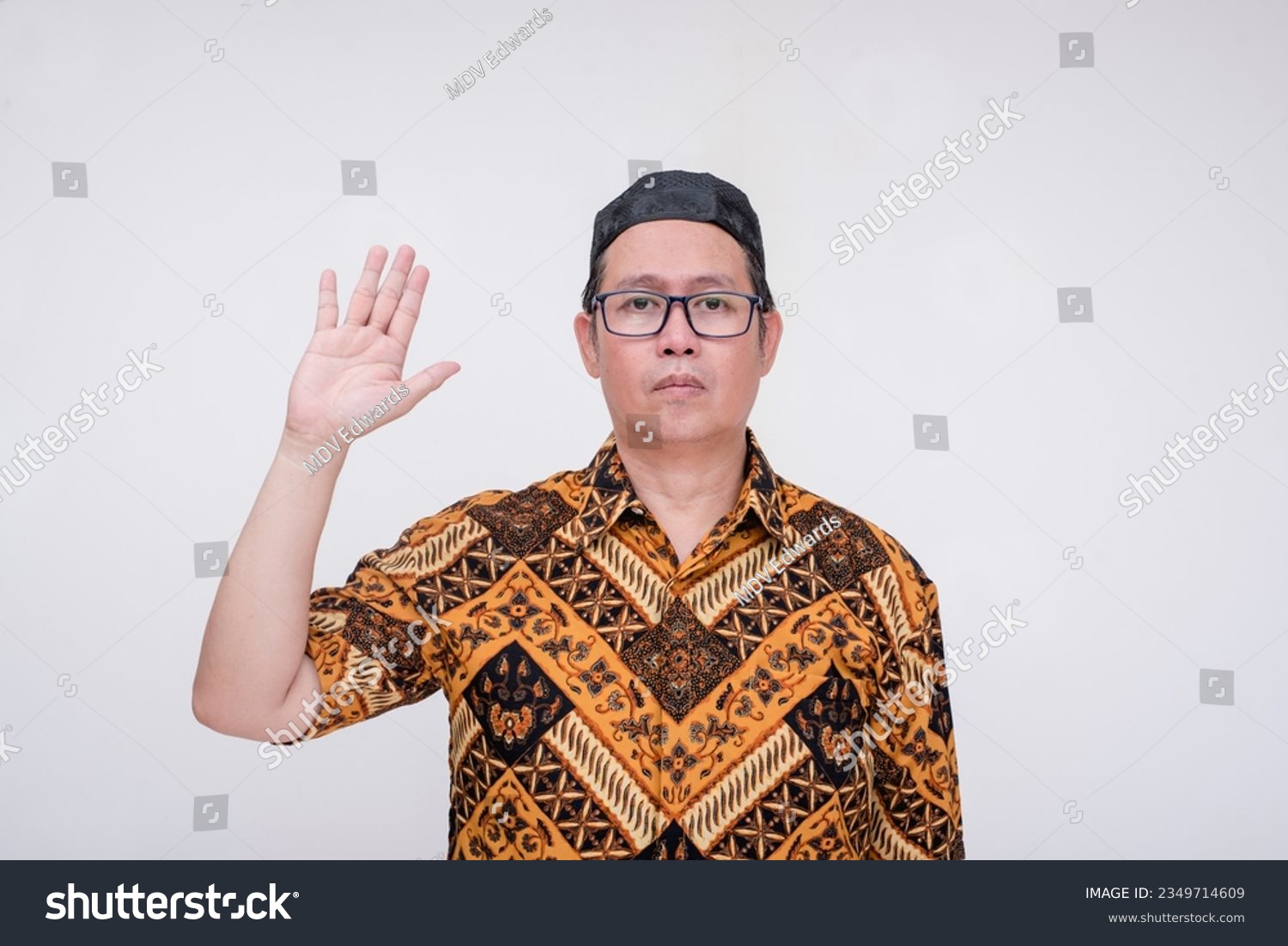 A southeast asian man pledges loyalty and makes an oath. Wearing a batik shirt and songkok skull cap with hand on chest. A middle aged muslim male in his 40s. Isolated on a white background. #2349714609