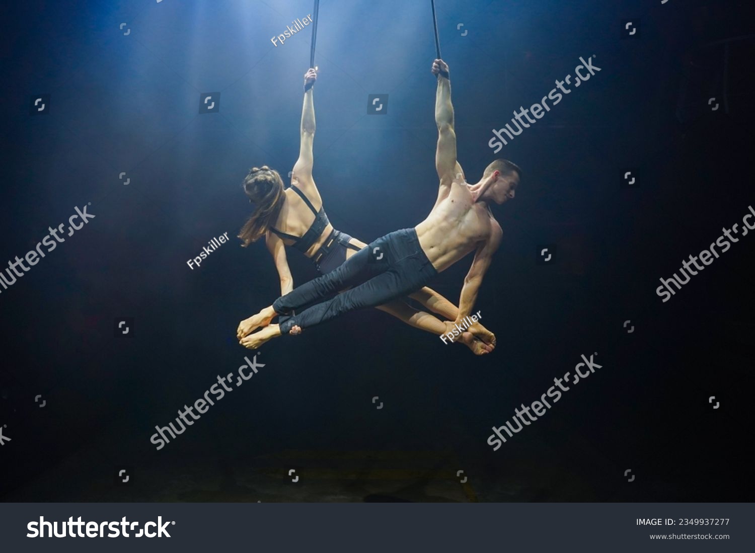 Aerial straps duo performance: a man and woman execute graceful acrobatic feats in mid-air against a black background, dressed in black and illuminated by a white-blue glow #2349937277