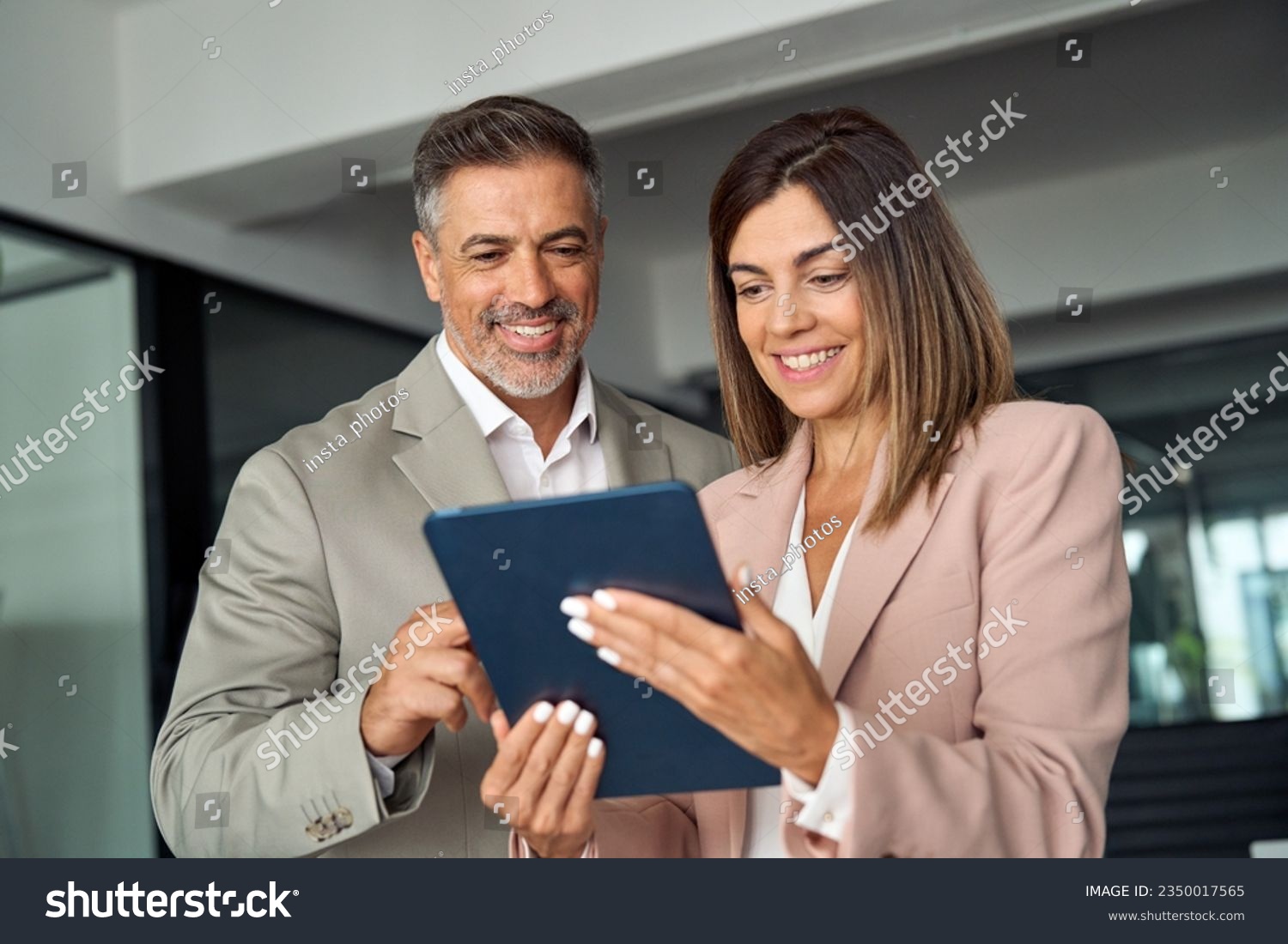 Smiling busy middle aged business man and business woman professional corporate executive leaders wearing suits discussing digital strategy using tablet computer standing in office working together. #2350017565