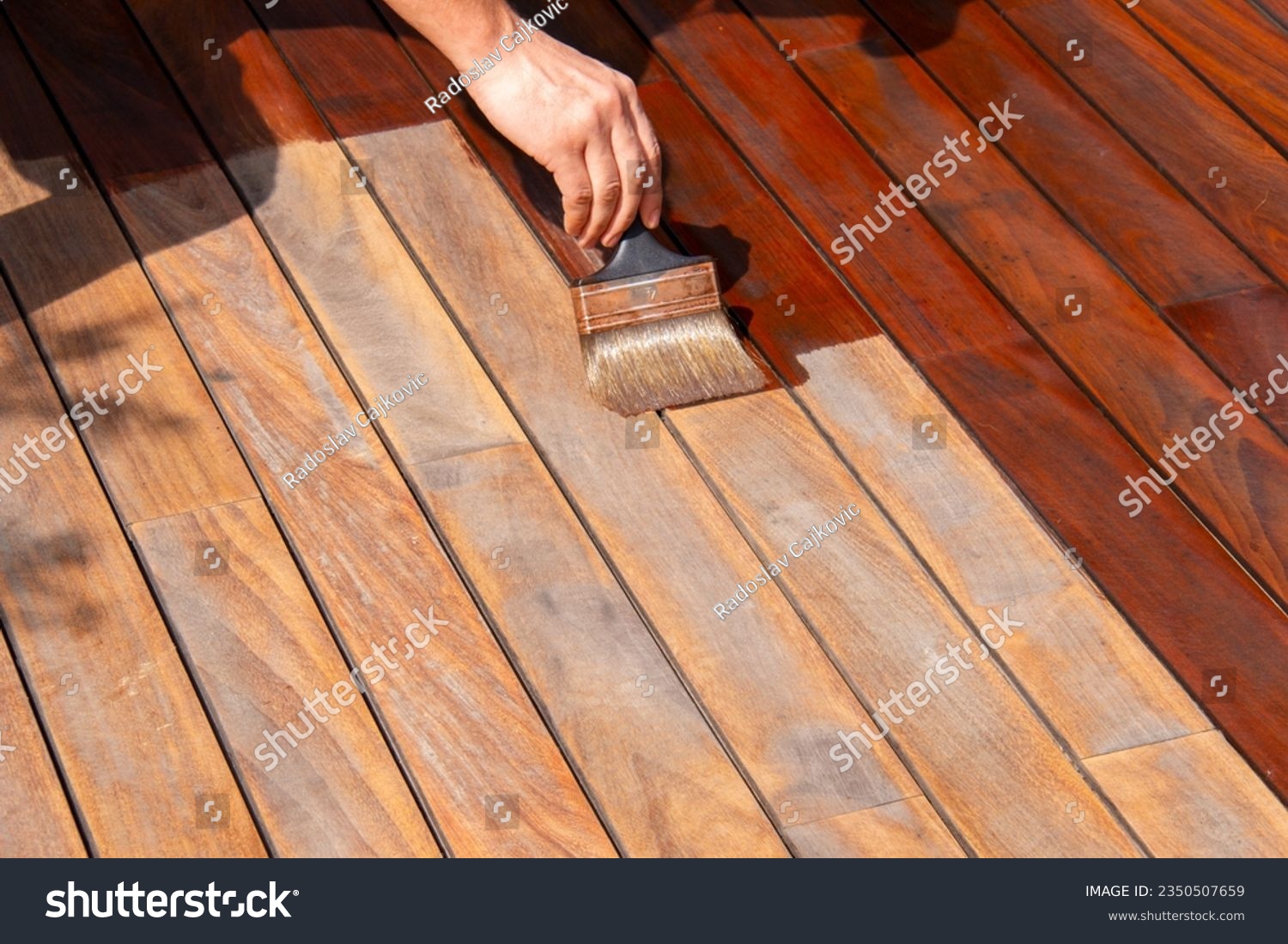 Woodworker refreshing wooden floor, worker's hand is applying decking oil on deck with a painting brush after sanding #2350507659