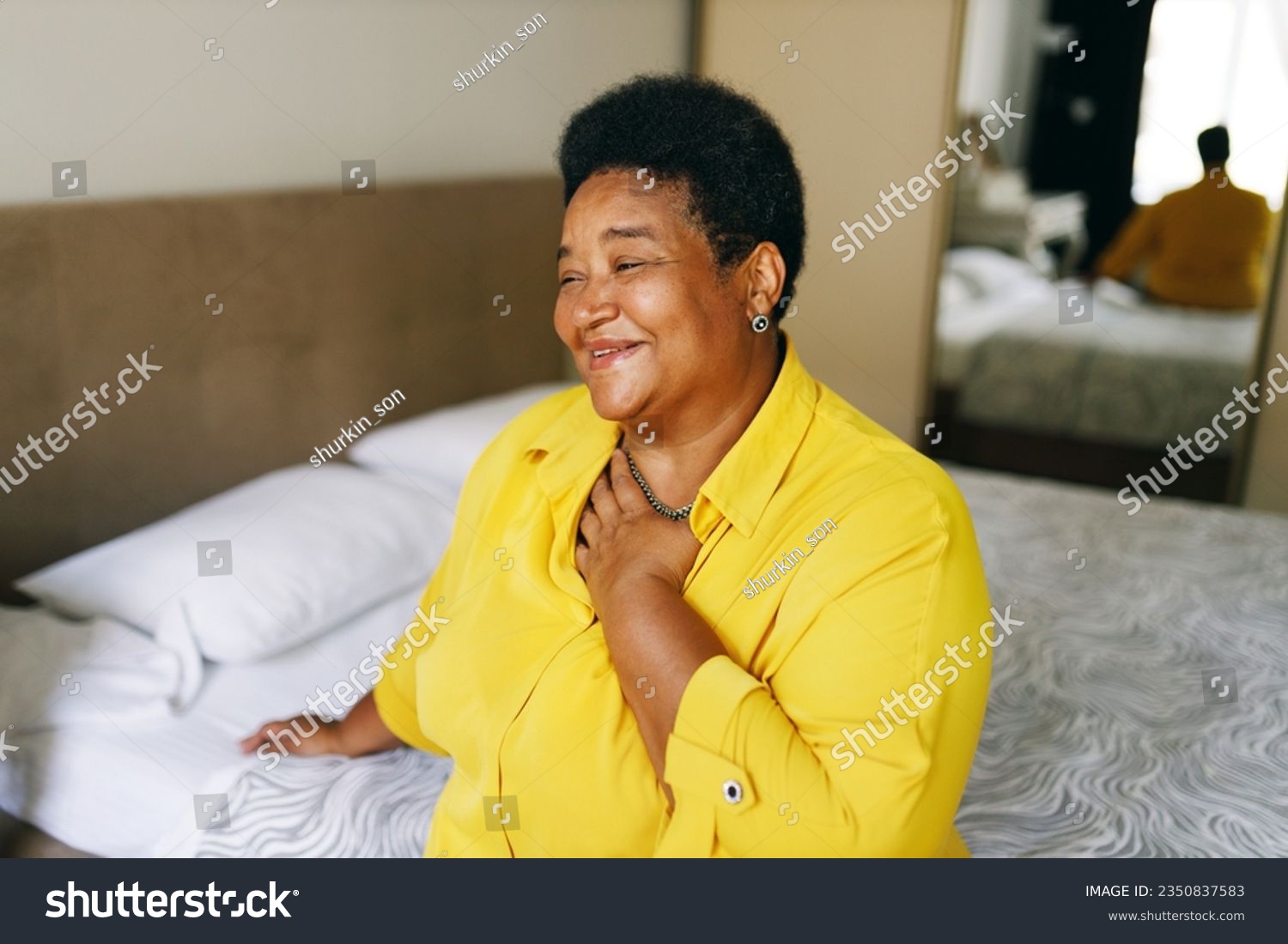 Happy cheerful grateful african american grandmother sitting on bed in yellow shirt keeping hand on chest smiling, being thankful for best life, thinking about her past, funny unforgettable moments #2350837583