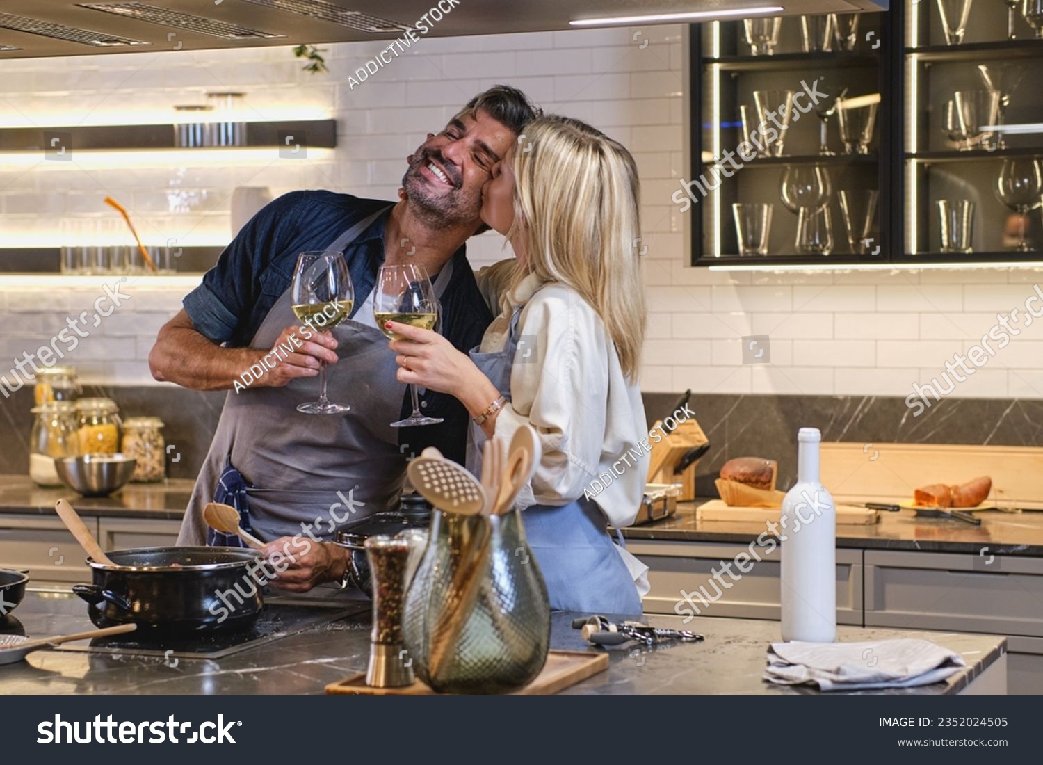 Loving wife kissing husband on cheek while cooking together at home and standing with glasses of white wine at stove #2352024505