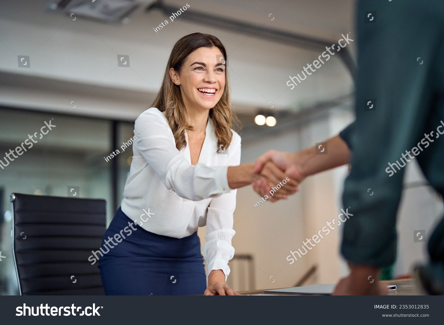 Happy mid aged business woman manager handshaking greeting client in office. Smiling female executive making successful deal with partner shaking hand at work standing at meeting table. #2353012835