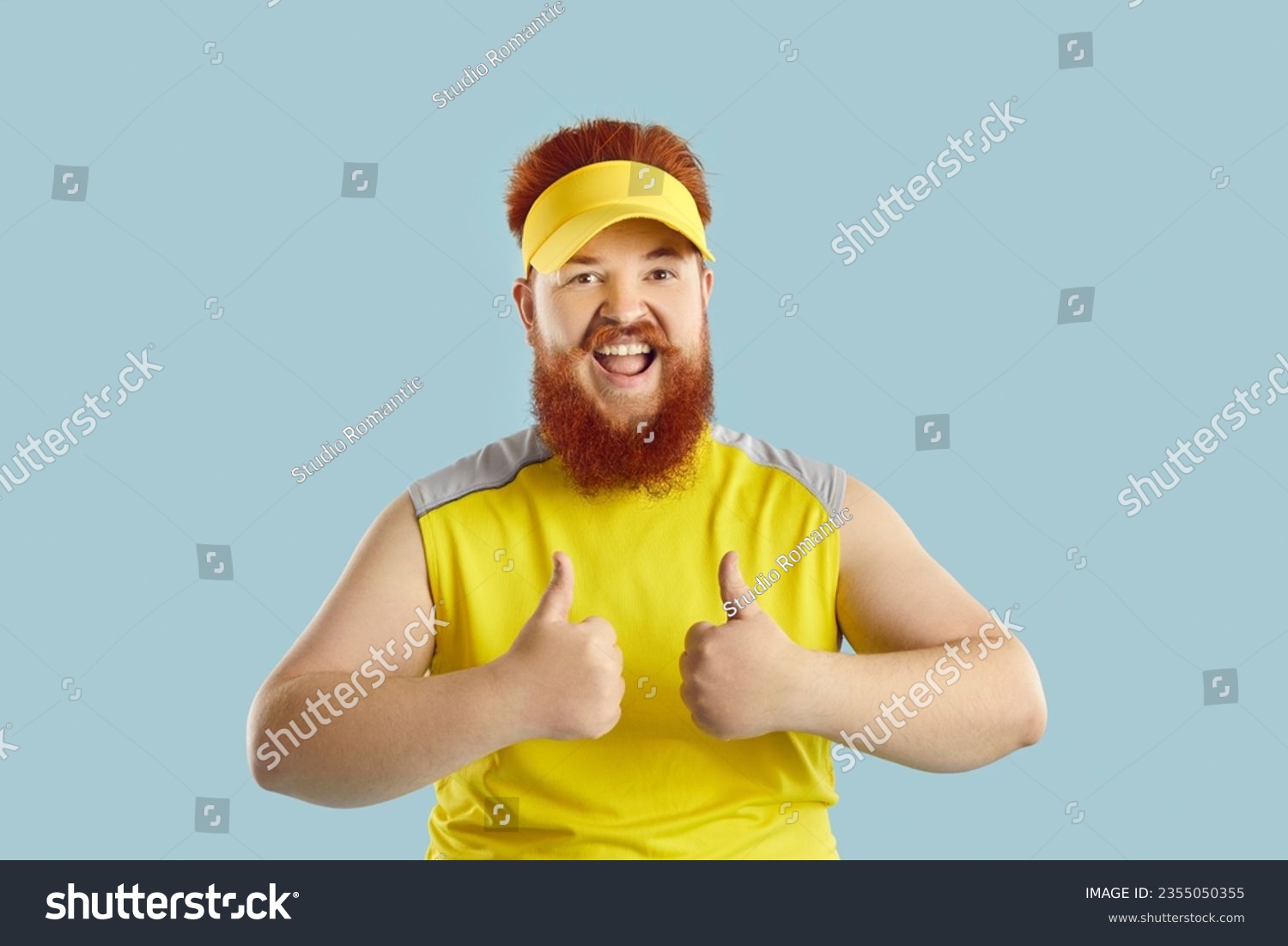 Funny happy fat man showing thumbs up motivating you to exercise isolated on light blue background. Young red-haired bearded man in sporty visor and T-shirt looks at camera with funny expression #2355050355