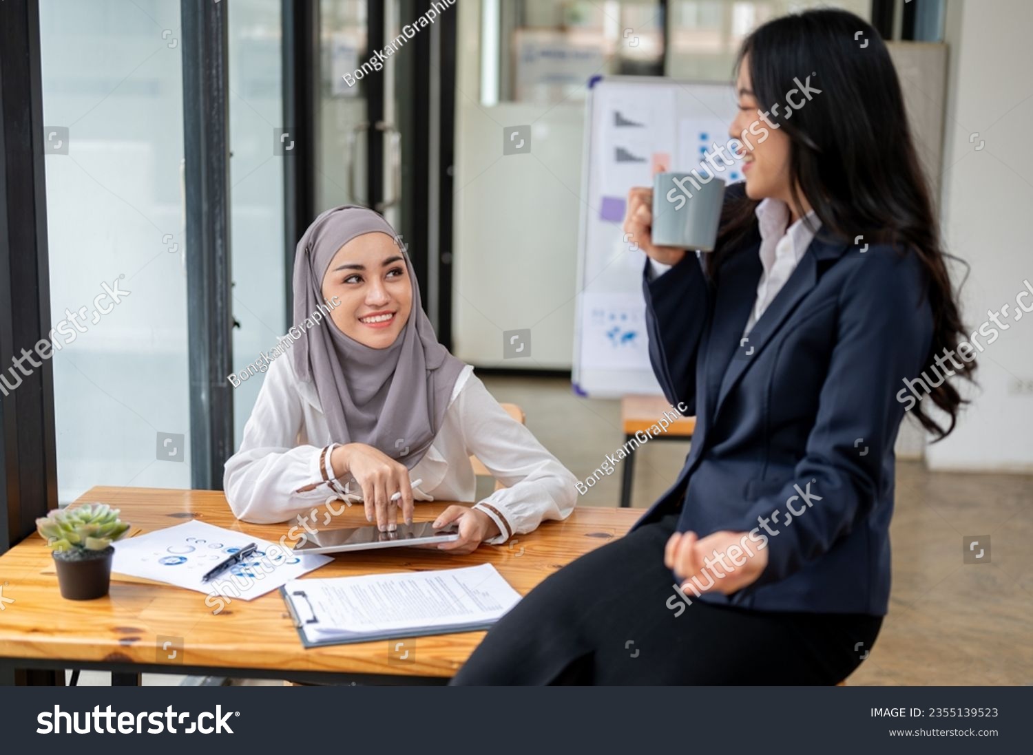 An attractive and smiling Asian Muslim female office worker wearing a hijab enjoys chatting with her colleague during a coffee break in the office. #2355139523