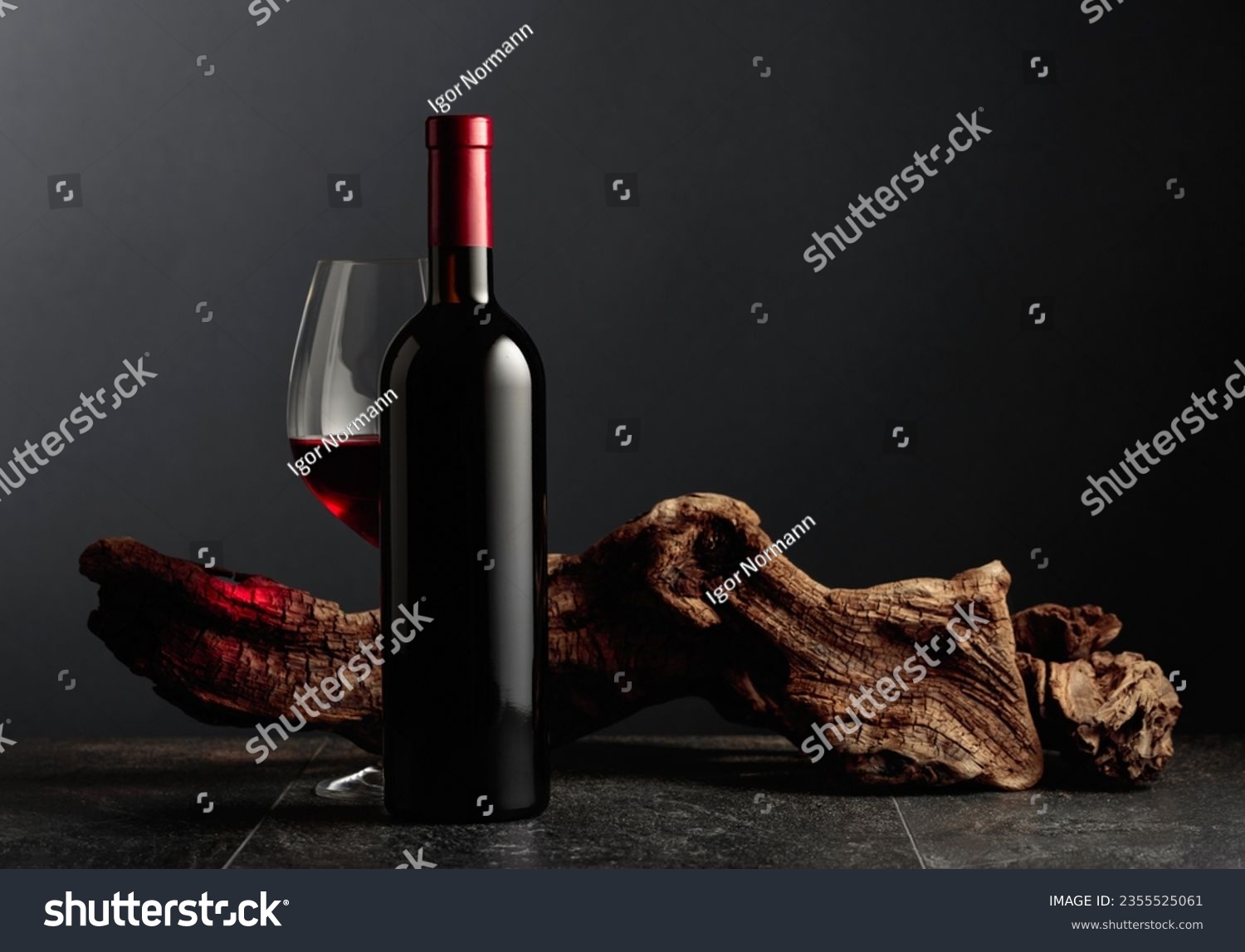 Bottle and glass of red wine on a black stone table. In the background old weathered snag. Frontal view with copy space. #2355525061