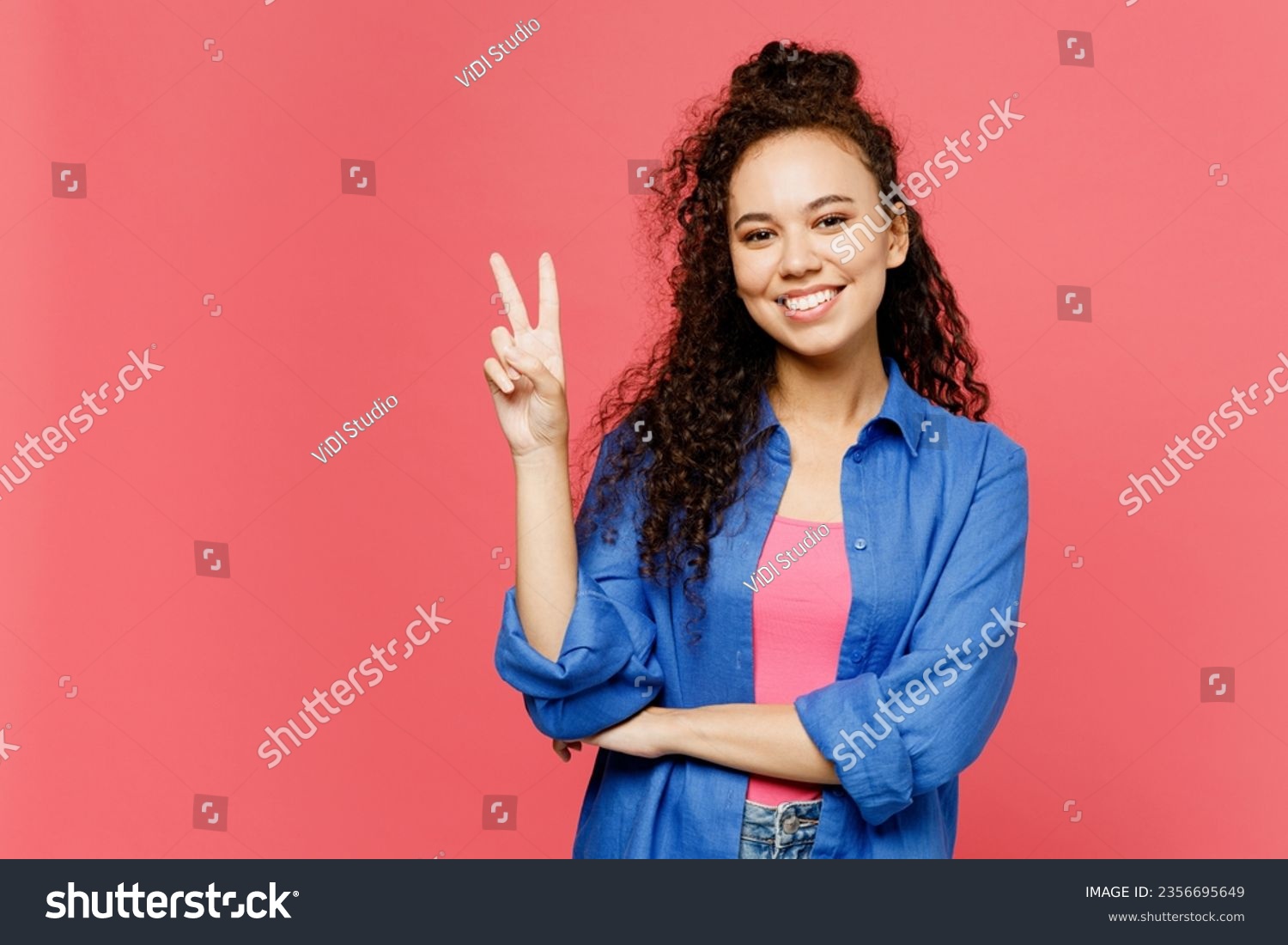 Young smiling happy cheerful woman of African American ethnicity she wear blue shirt casual clothes showing victory sign look camera isolated on plain pastel pink background studio. Lifestyle concept #2356695649