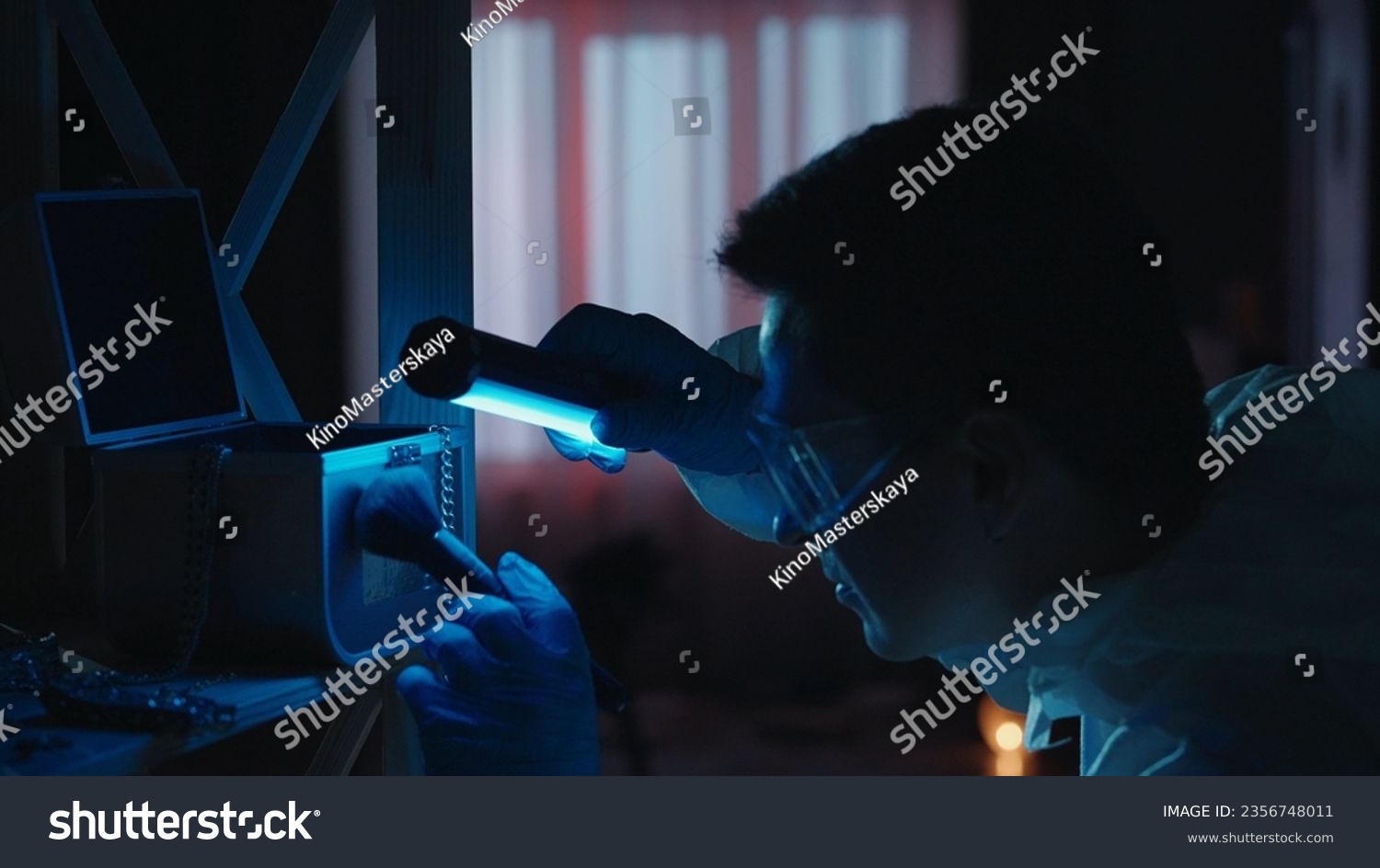 A male expert criminologist removes fingerprints from a jewelry box with a brush. A man gathers evidence using an ultraviolet lamp at a crime scene in a dark apartment lit by red, blue police sirens. #2356748011