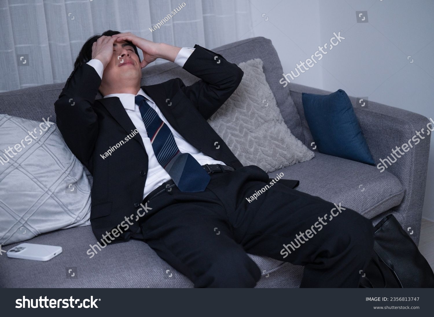 A Japanese male office worker, tired from a late-night workday, lounging on his sofa after returning home. #2356813747