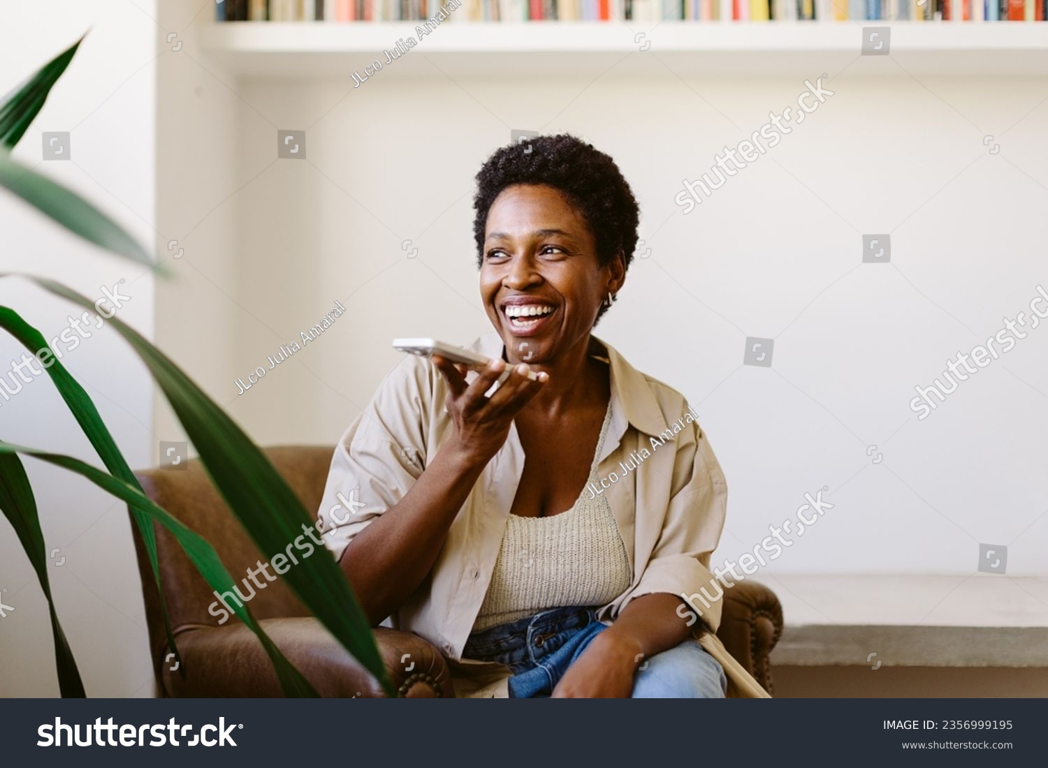 Happy Brazilian woman sits on a couch, speaking on her smartphone. Smiling and relaxed, she holds the phone and looks away, fully engaged in a phone call from her living room. #2356999195