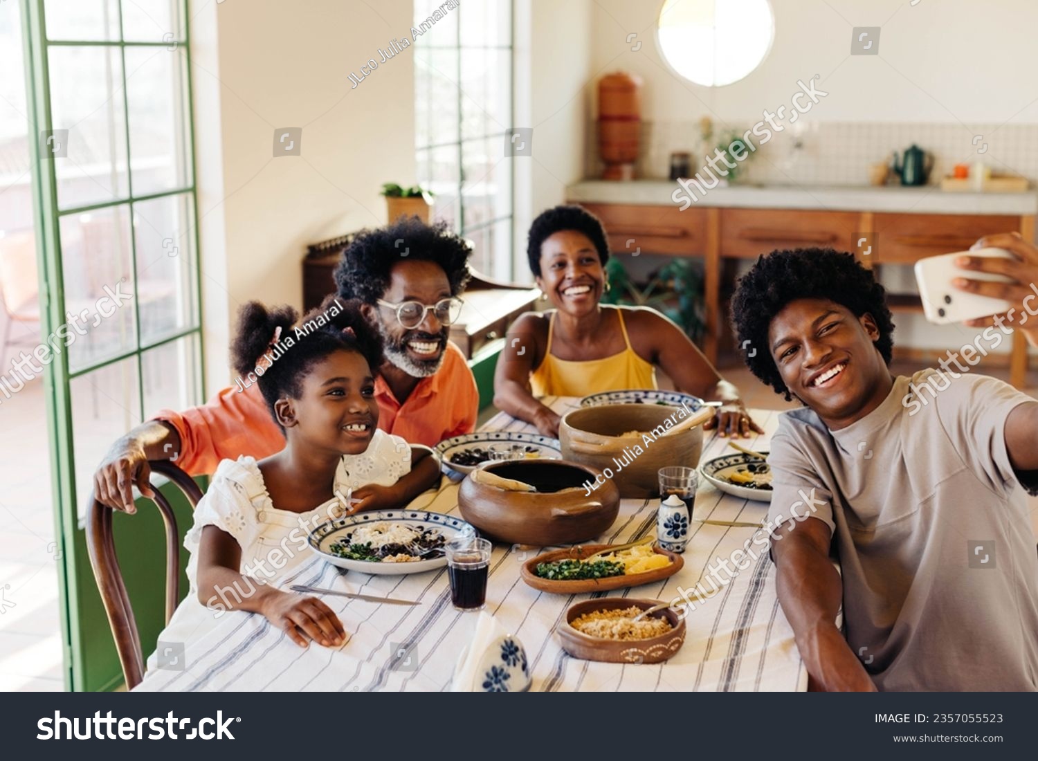 Happy Brazilian family sitting at the table for a traditional meal, with clay pots filled with delicious dishes. They smile and pose for a selfie, creating memories in their kitchen. #2357055523