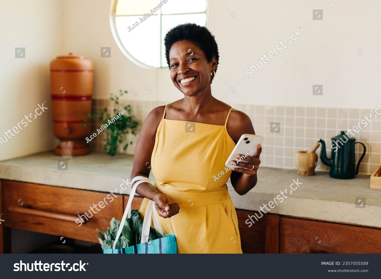 Smiling Brazilian woman stands in her kitchen, holding a bag of fresh vegetables. She confidently poses with her mobile phone, capturing the essence of her cheerful cooking and happy lifestyle. #2357055589