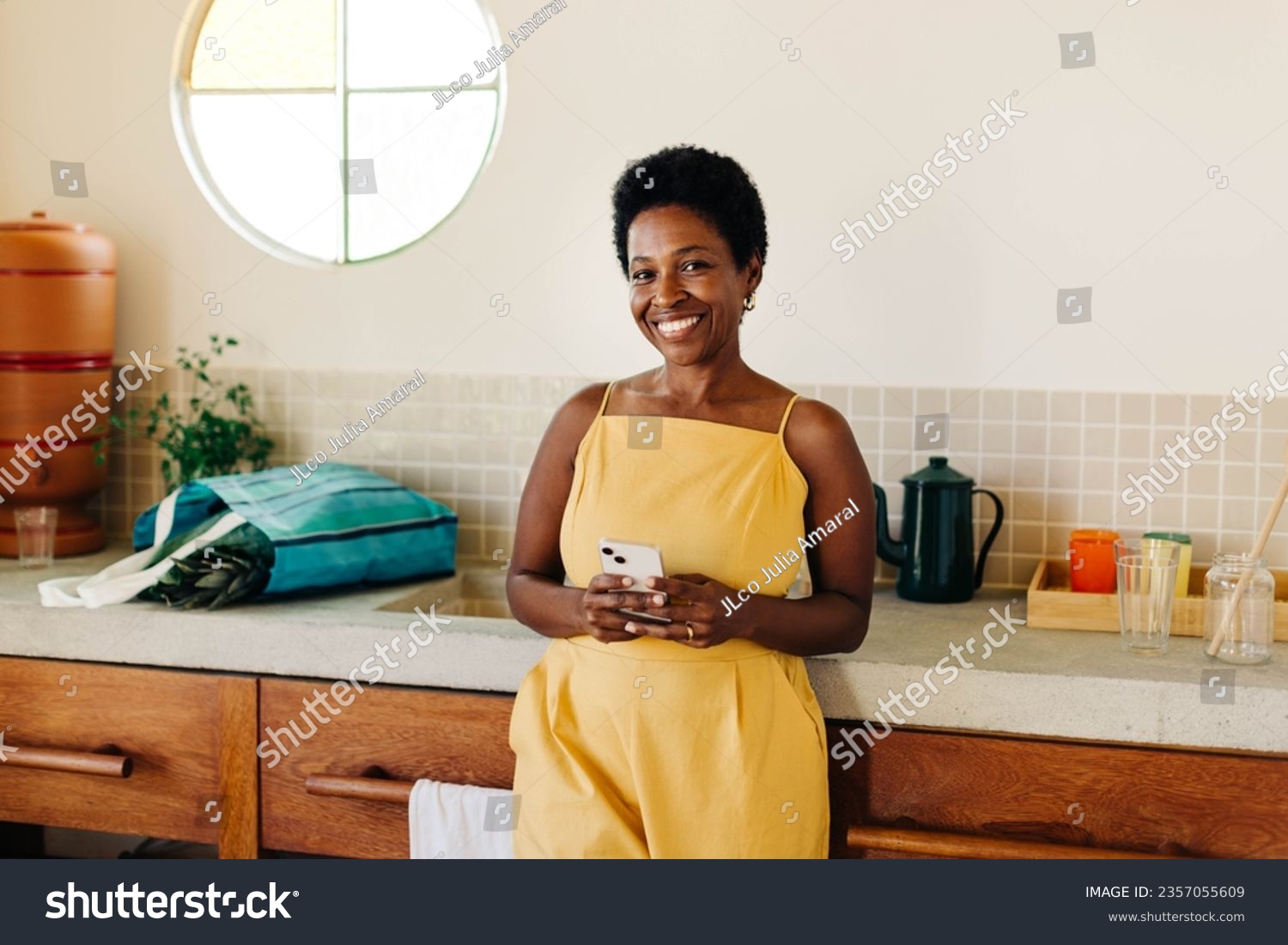 A happy Brazilian woman in a kitchen, using her mobile phone with a cheerful smile. #2357055609