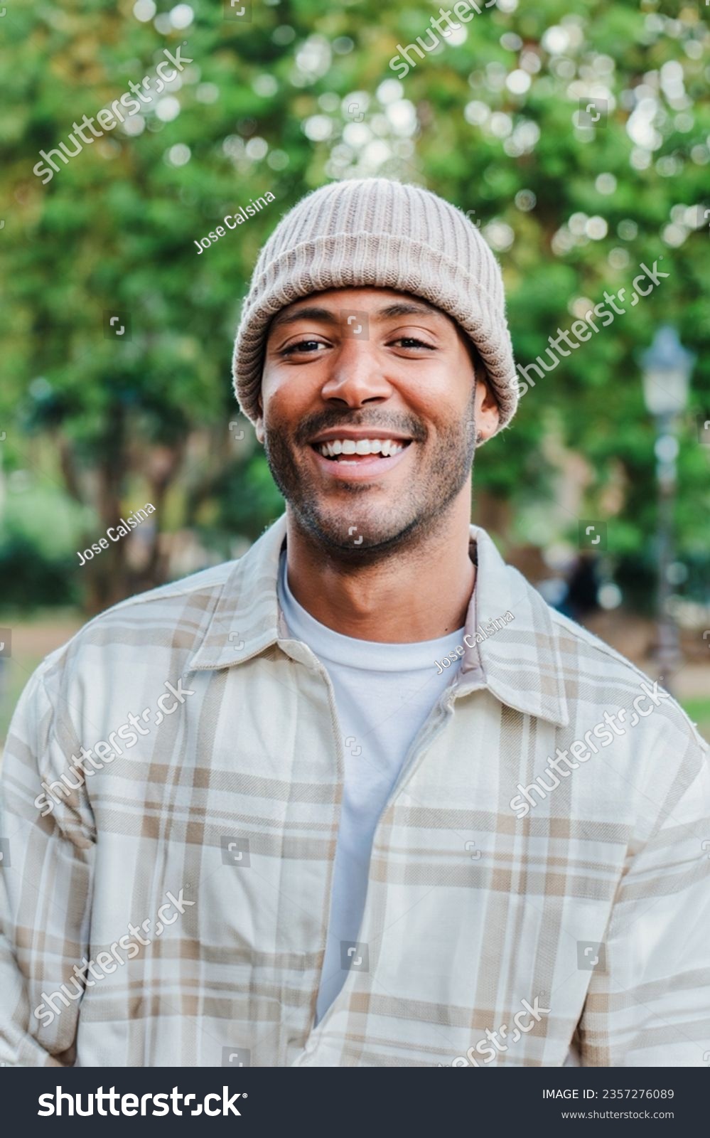Vertical close up portrait of young hispanic man with a beanie hat smiling and looking at camera outdoors. Front view of latin happy guy standing at park with carefree attitude. Lifestyle concept #2357276089