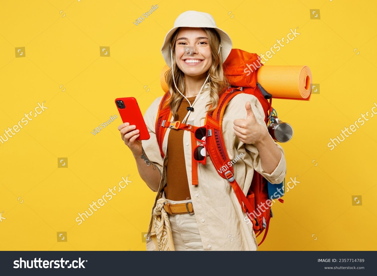 Young woman carry backpack with stuff mat use mobile cell phone show thumb up isolated on plain yellow background Tourist leads active lifestyle walk on spare time Hiking trek rest travel trip concept #2357714789