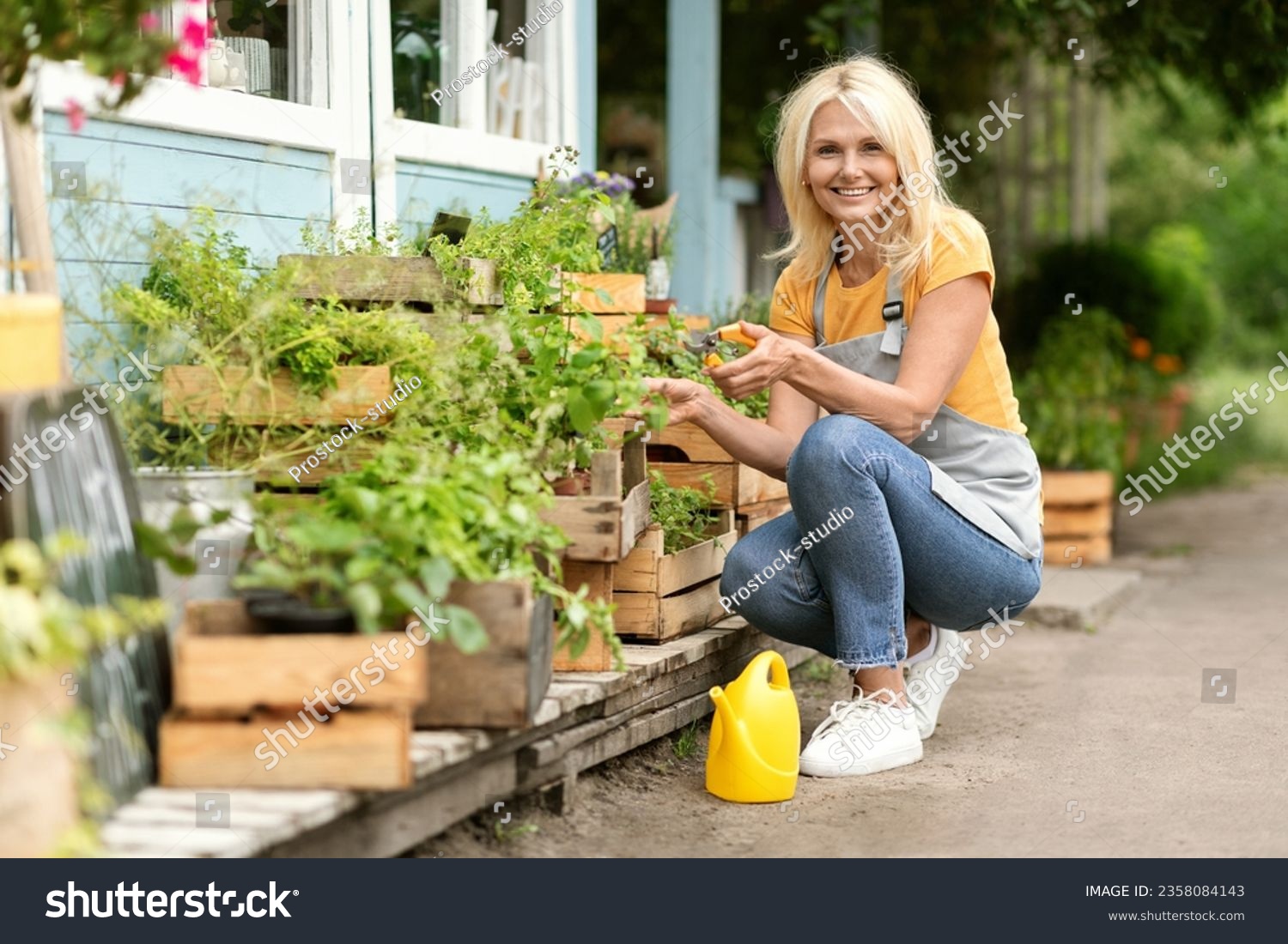 Smiling Mature Woman Taking Care About Plants In Crates At Her Backyard, Happy Beautiful Senior Lady Wearing Apron Enjoying Gardening And Eco Farming, Using Pruning Shears Secateurs, Free Space #2358084143