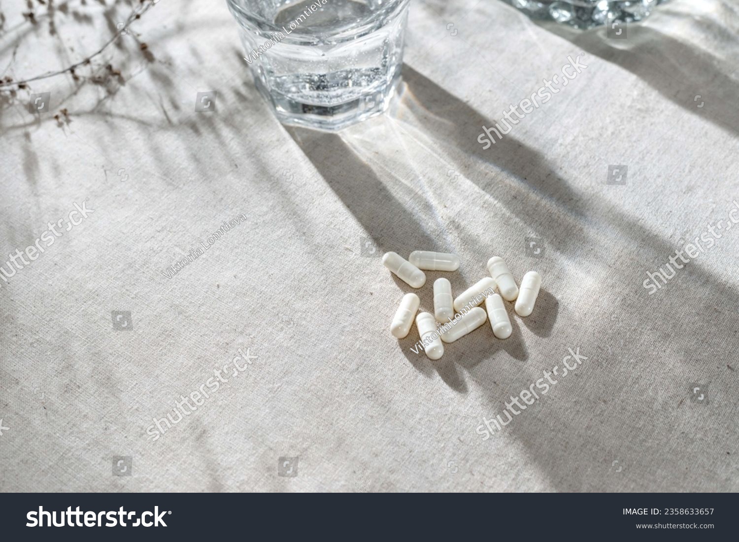 White capsules, glass with water and dried grass on neutral beige linen tablecloth with aesthetic light shadows. #2358633657