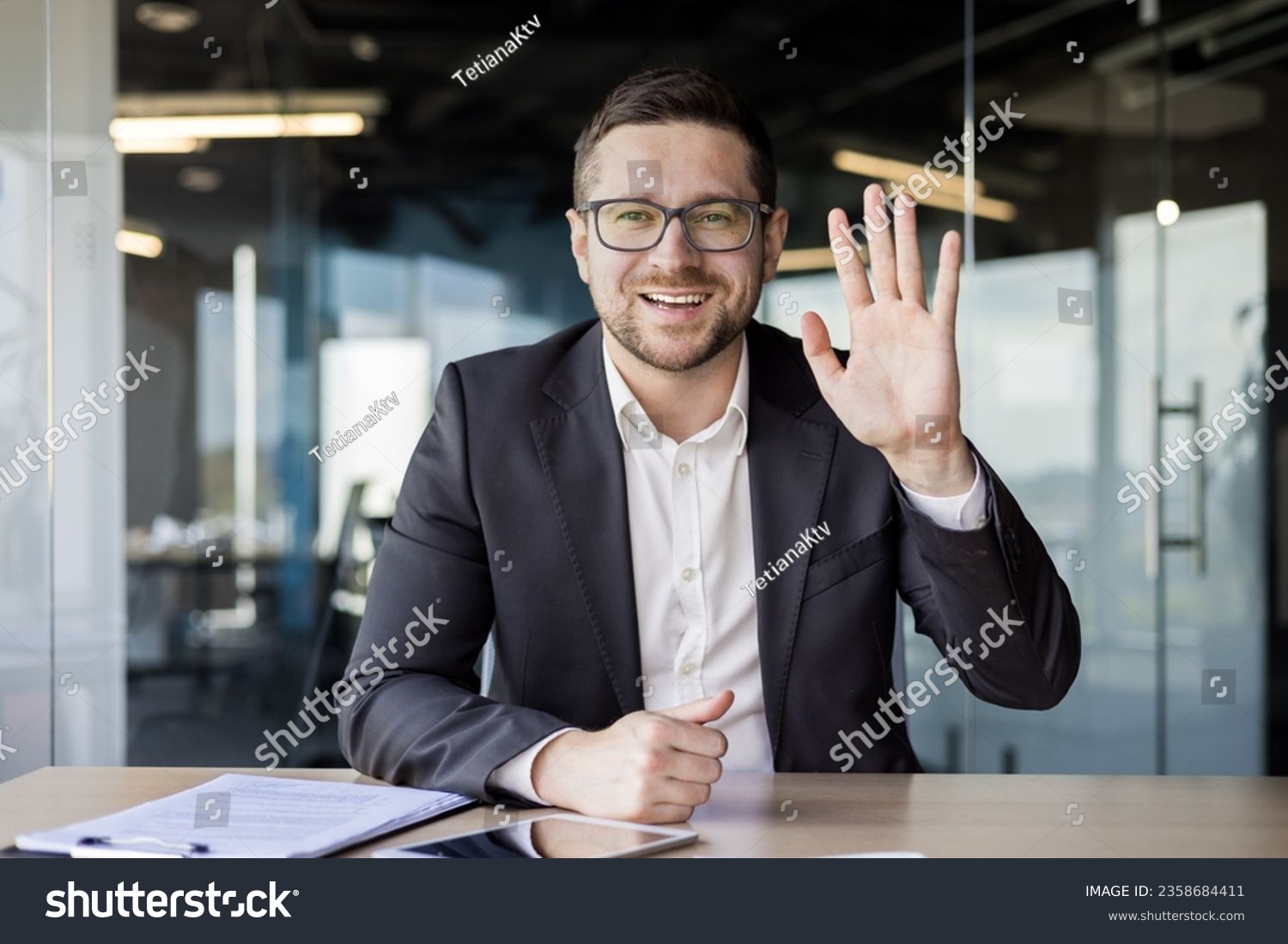 Portrait of a young businessman in a suit who sits in the office in front of the camera, communicates online, greets with his hand. #2358684411