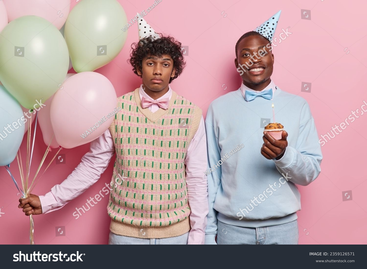 Horizontal shot of curly haired Hindu man and black man dressed in festive clothing holds cupcake with candle holds bunch of colorful balloons celebrates special occasion isolated over pink background #2359126571