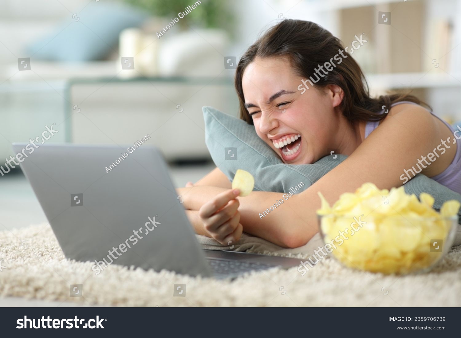 Funny woman eating potato chips watching media on laptop lying on the floor laughing hilariously at home #2359706739
